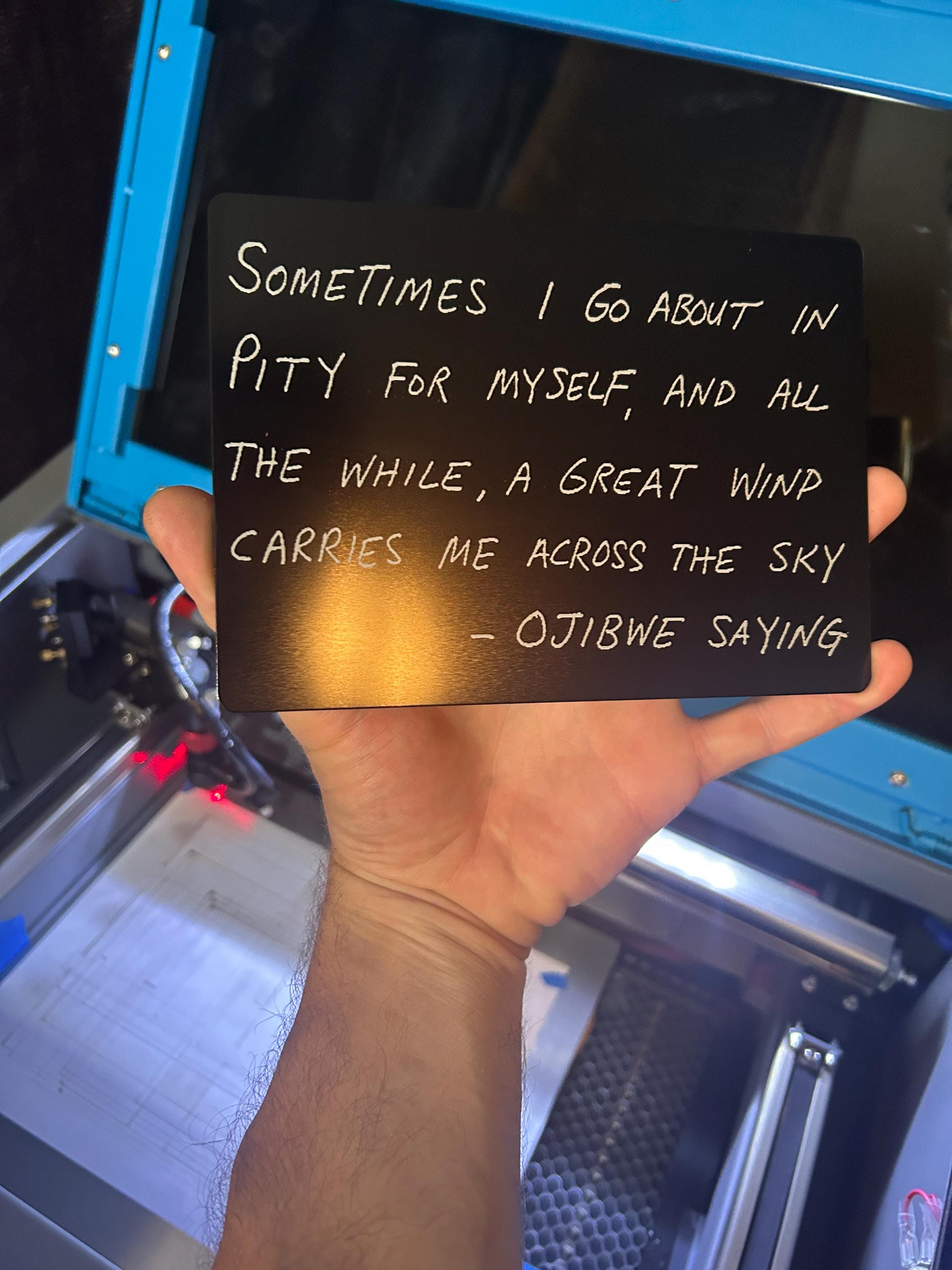 Hand holding a black sign with text in front of a laser machine
