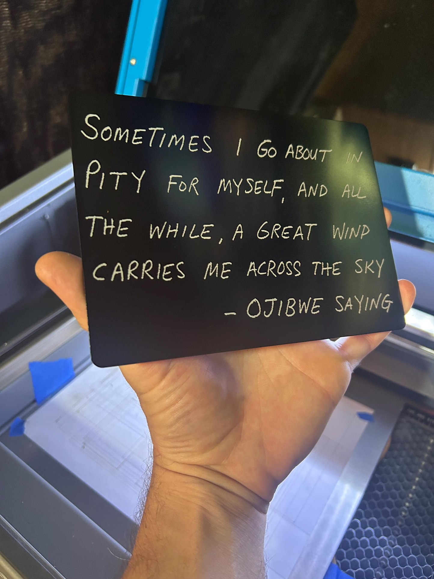 Hand holding a black and silver laser engraved ojibwe quote plaque with inscribed quote against a blurred background.