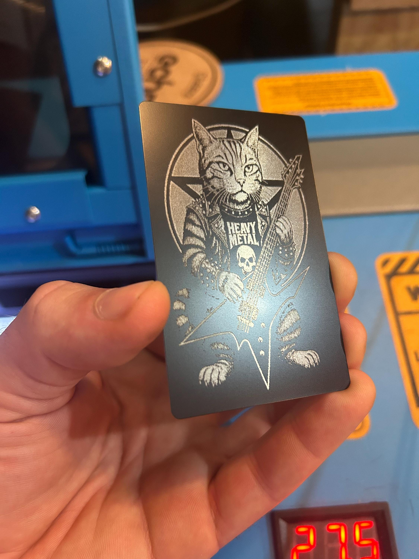 Hand holding a card with a cat playing guitar and heavy metal illustration art, against a blue laser machine background.