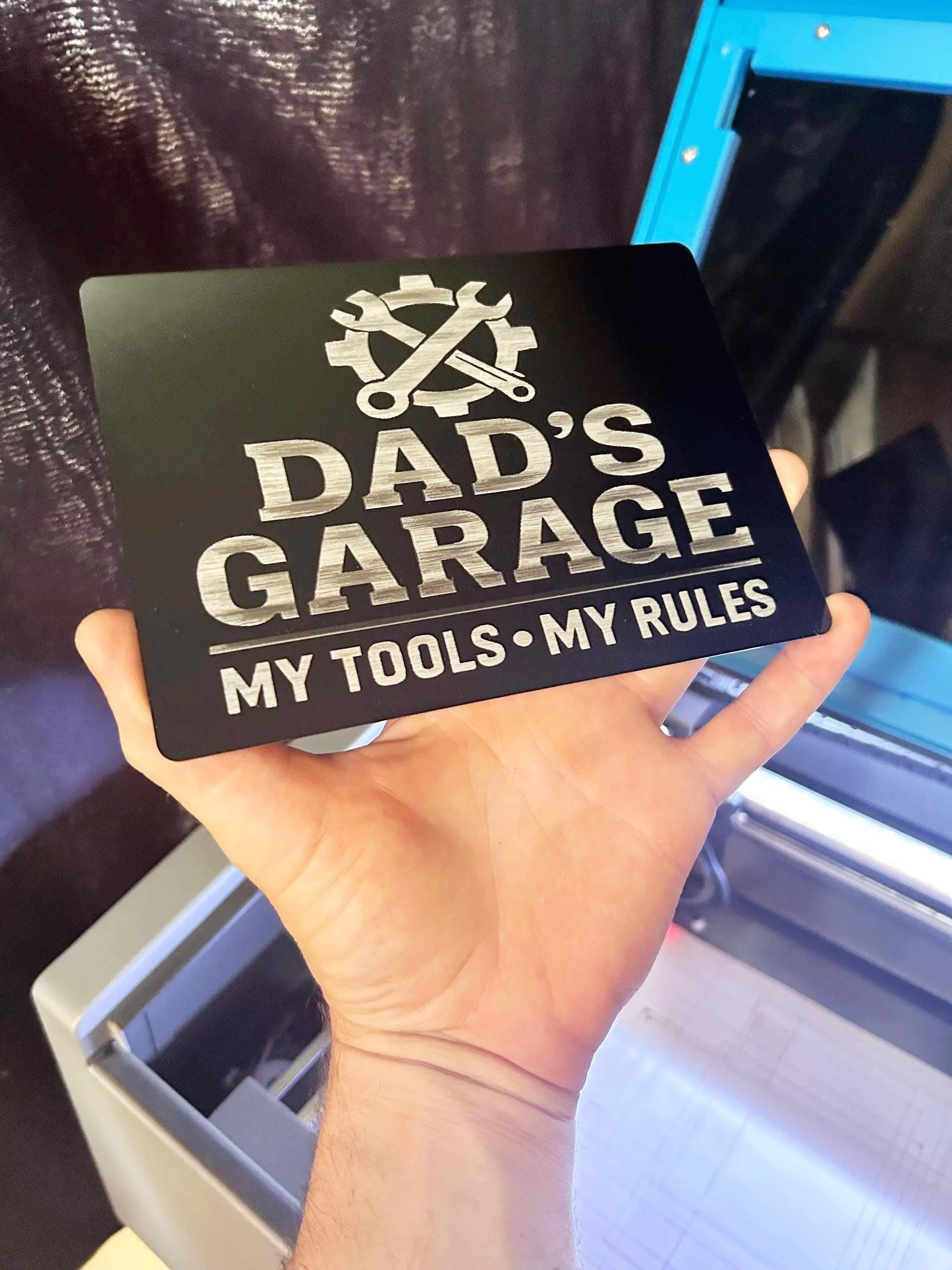 Hand holding a black/silver sign with 'Dad's Garage, My Tools, My Rules' text.
