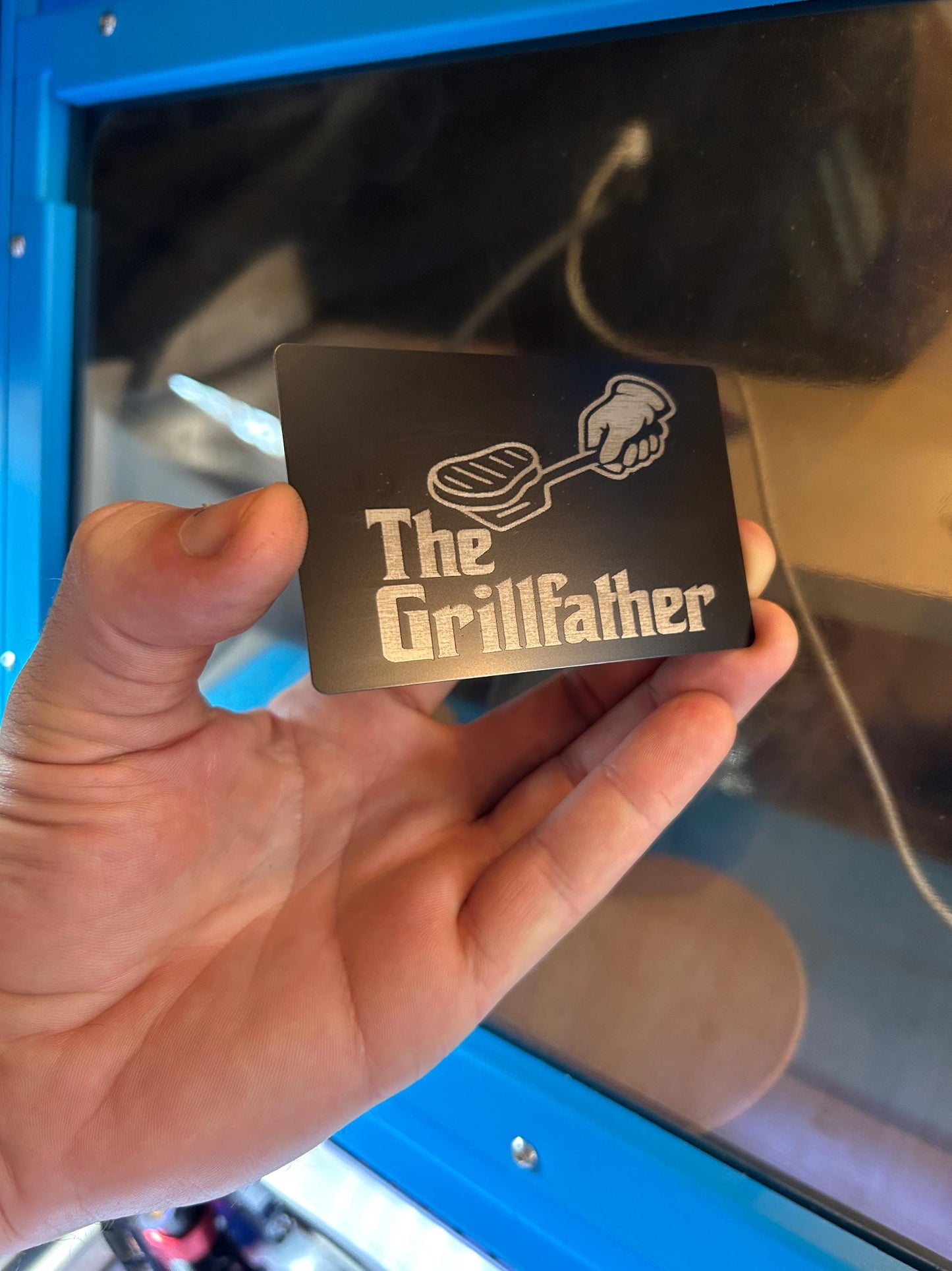 Hand holding a metal father's day gift card with 'The Grillfather' design in front of a blue machine.