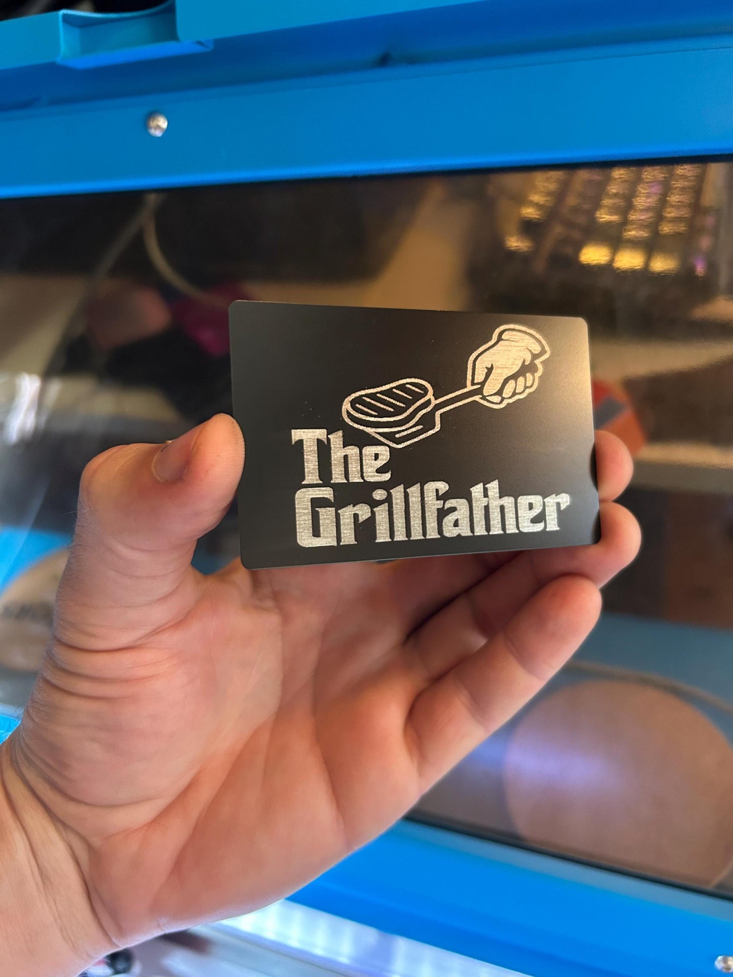 Hand holding a metal engraved card in black/silver with 'The Grillfather' design in front of a blue machine.