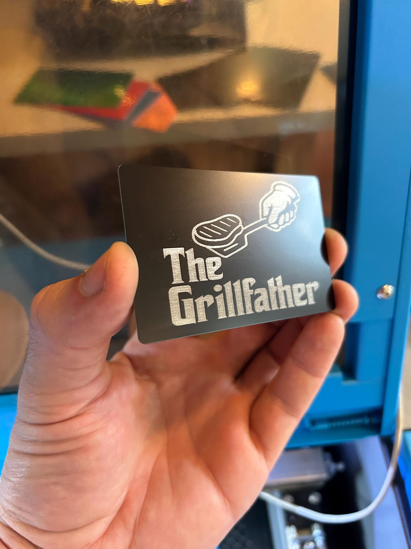 Hand holding a card with 'The Grillfather' design in front of a laser machine.