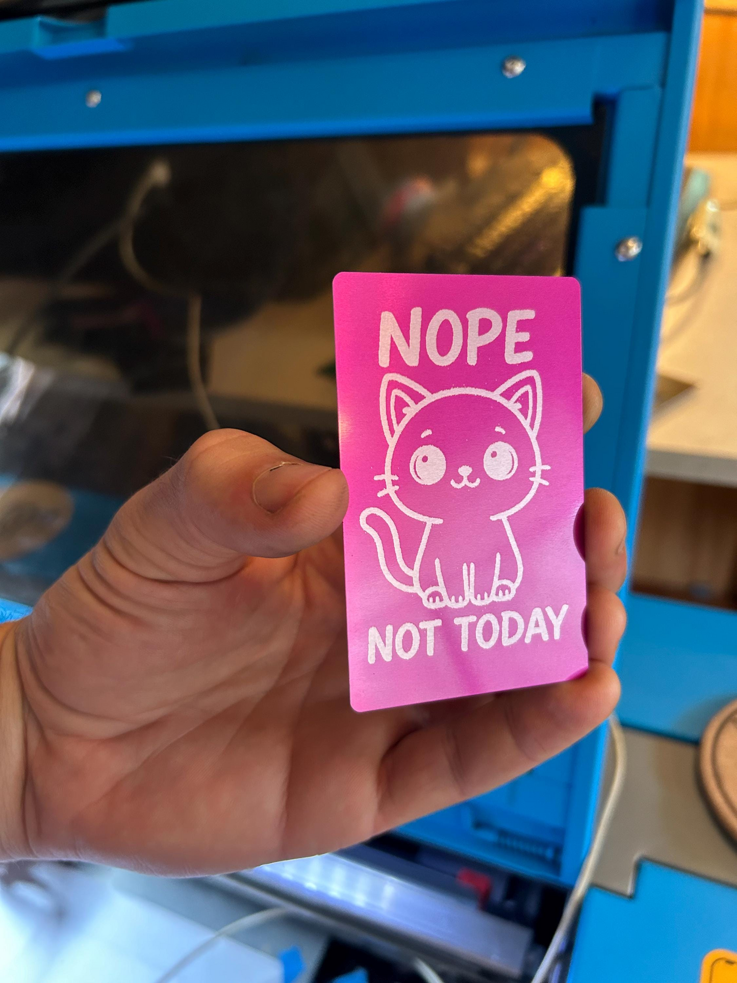 Pink metal keepsake card with a cat illustration and text that reads: Nope, Not Today -- held by a hand in front of a blue machine.