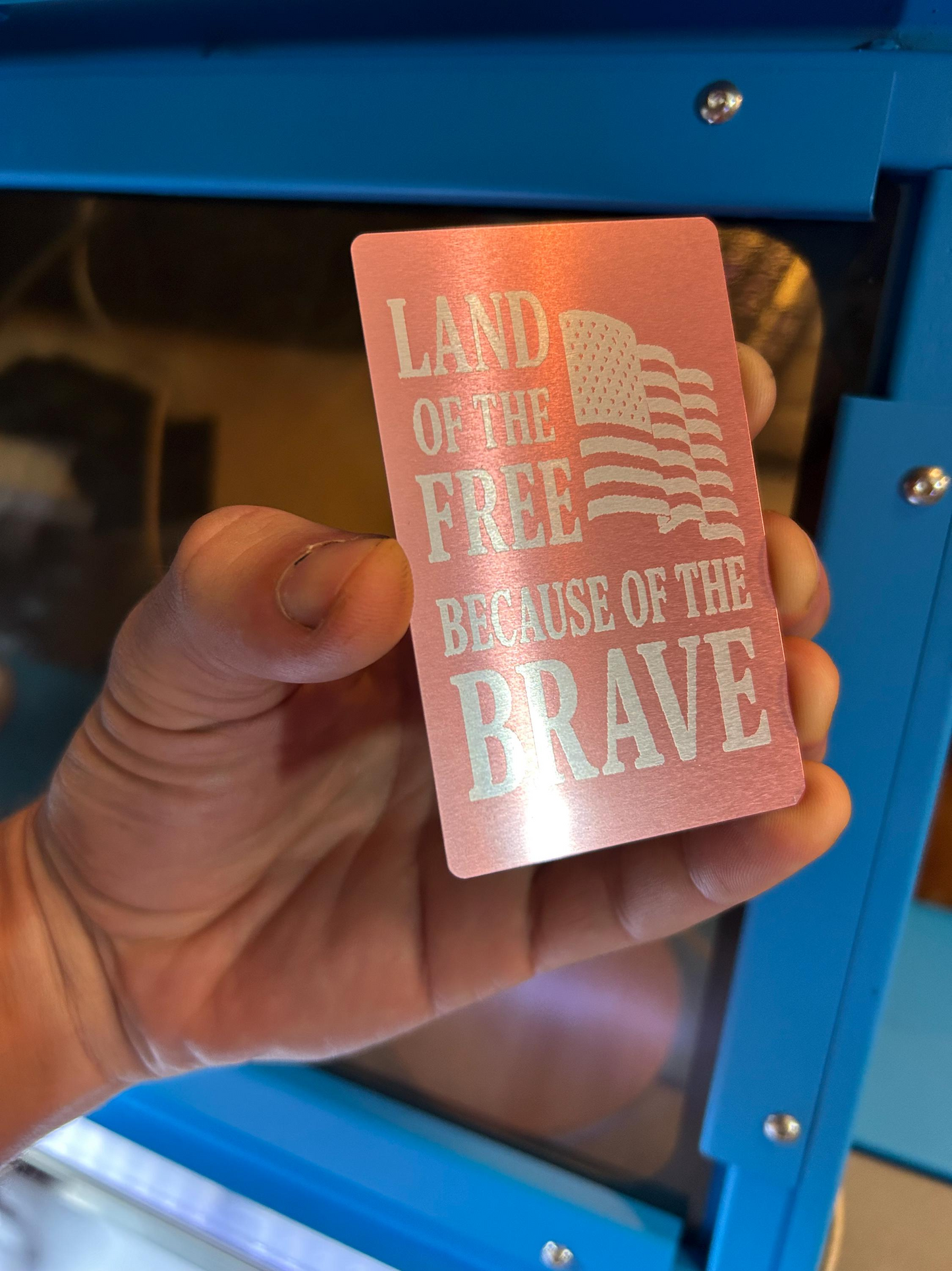Hand holding a laser engraved pink card with 'Land of the Free Because of the Brave' text and American flag design.