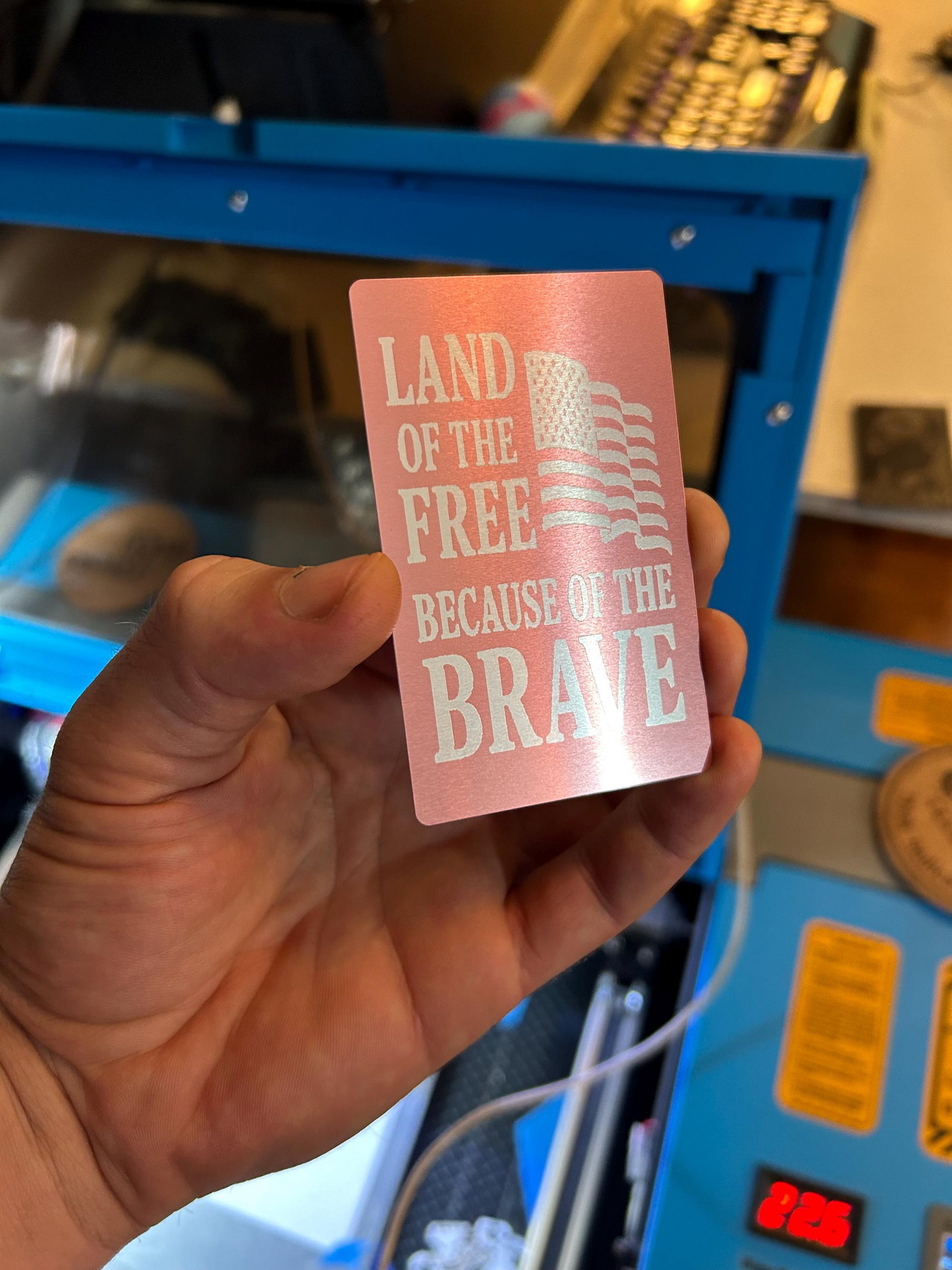 Hand holding a metal rose gold card with 'Land of the Free Because of the Brave' text and American flag design.