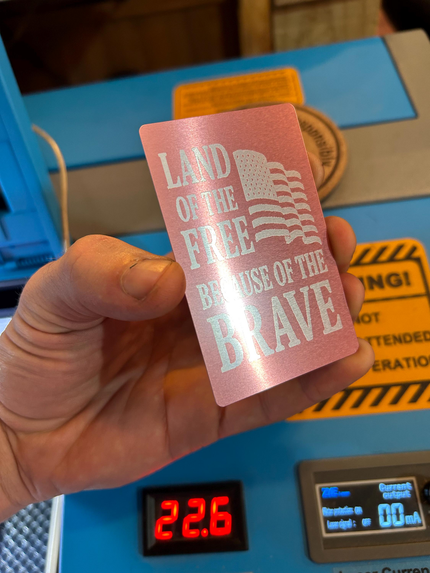 Hand holding a pink card with text and an American flag design, with a blue machine and digital display in the background.