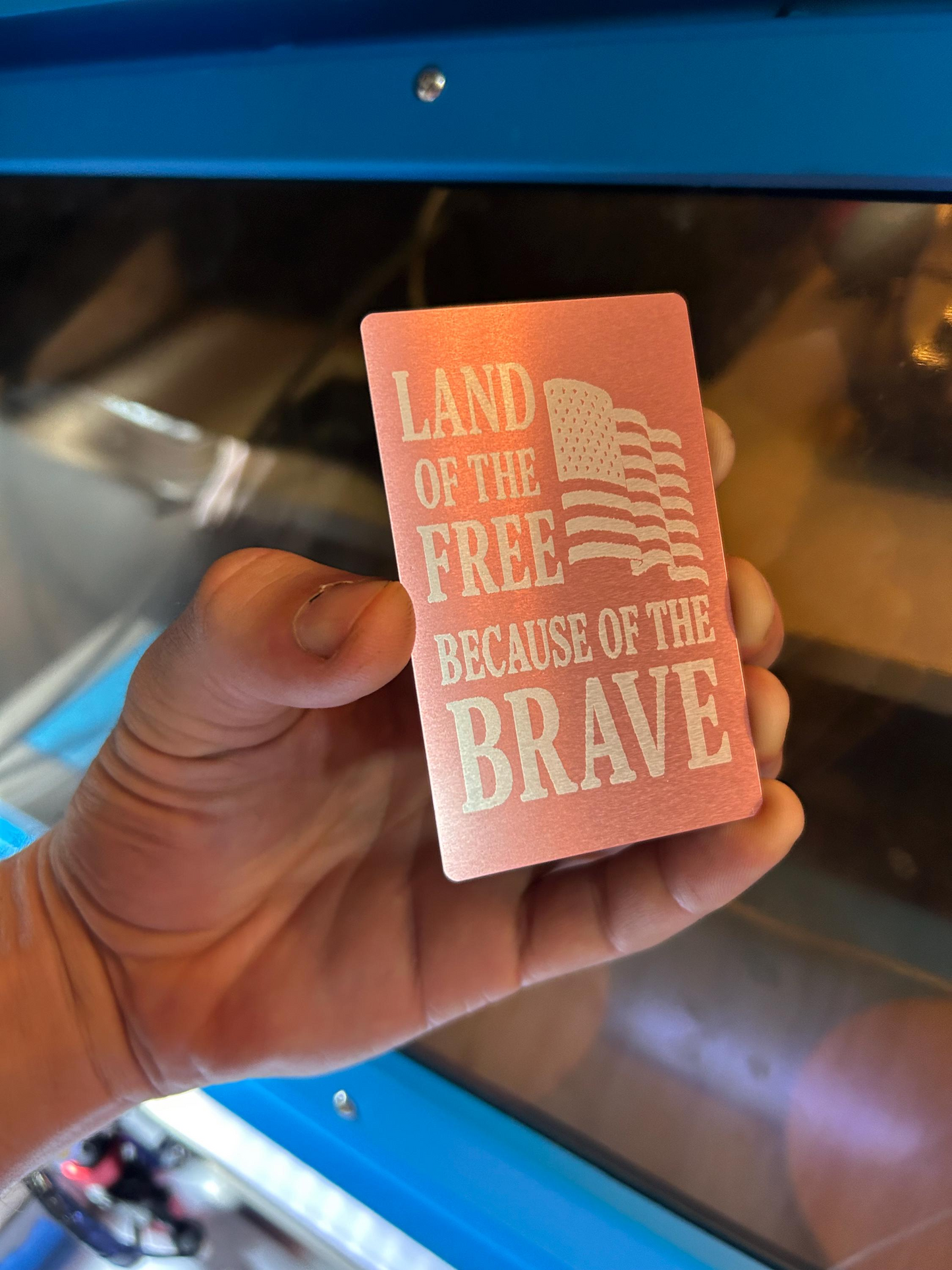 Hand holding a pink metal placard gift with 'Land of the Free Because of the Brave' text and American flag design.