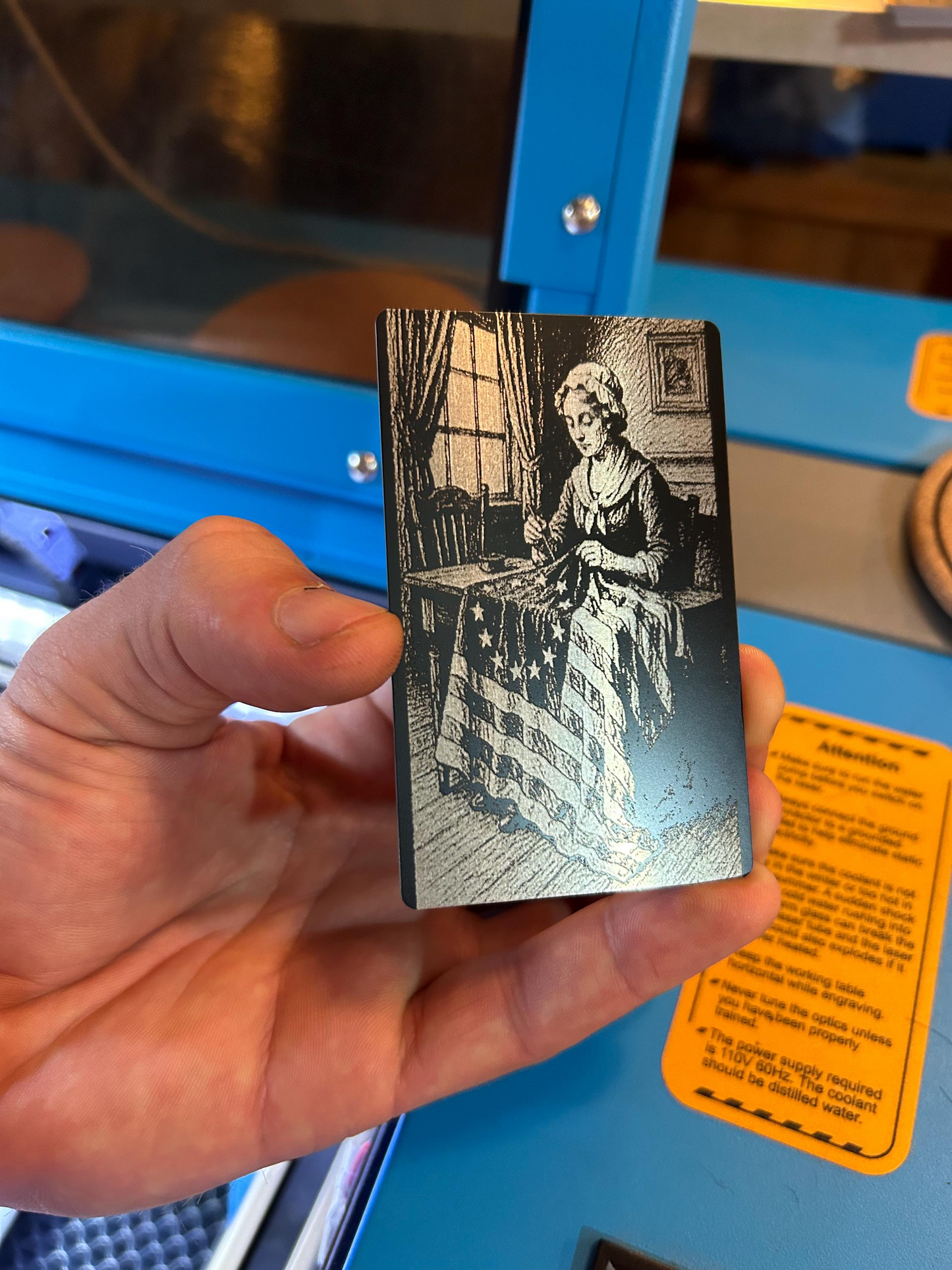 Hand holding a small engraved card with a detailed illustration of Betsy Ross and the USA flag in a room.