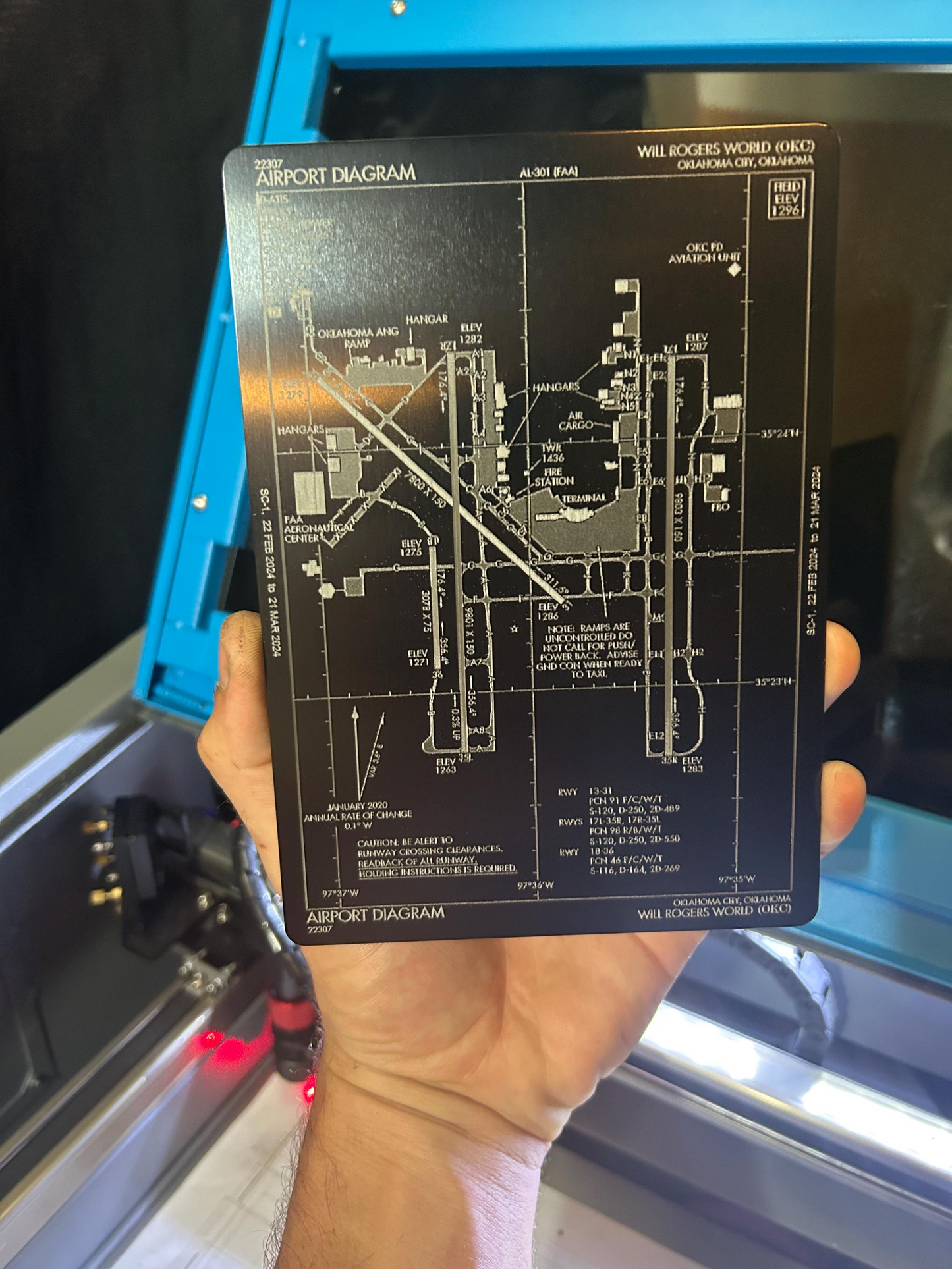 Hand holding a metal airport diagram placard in front of a blue machine.