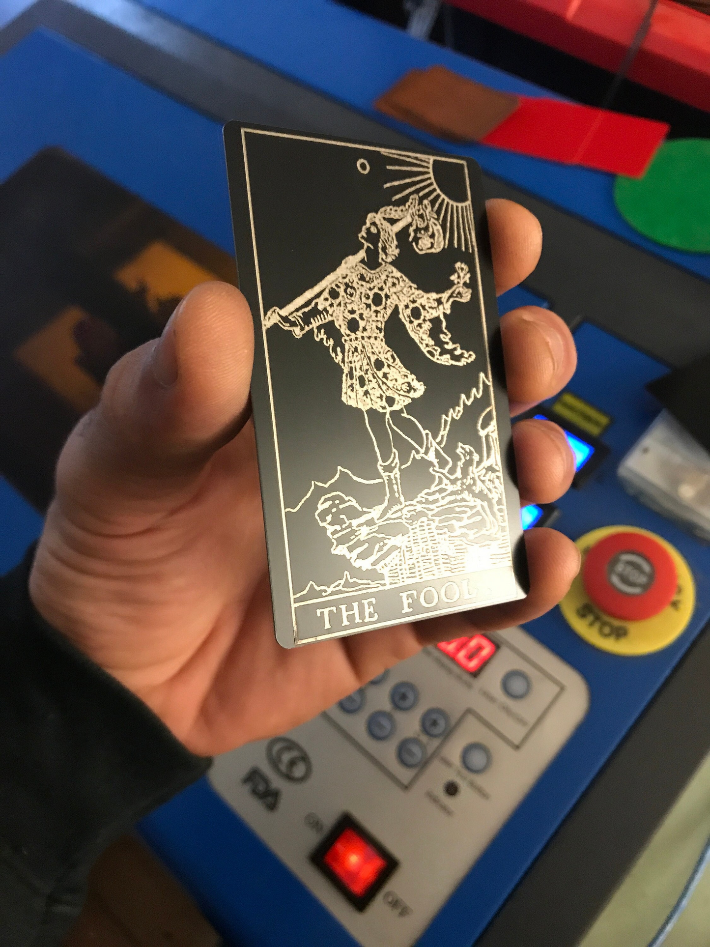 Hand holding a metal laser engraved tarot card with 'The Fool' symbol in front of a cnc laser machine.
