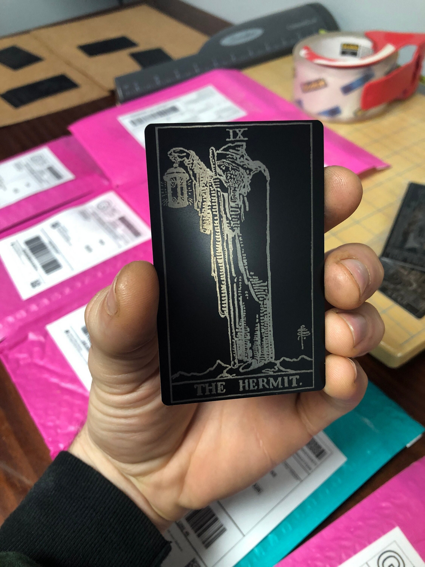 Hand holding a black metal laser engraved tarot card with white text and design, possibly a tarot card, on a background full of mailing packages