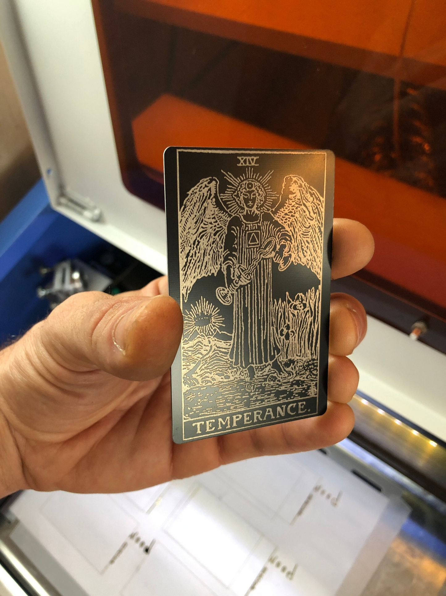 Hand holding a laser engraved black/silver metal tarot card with angel design and 'Temperance' text in front of a machine.
