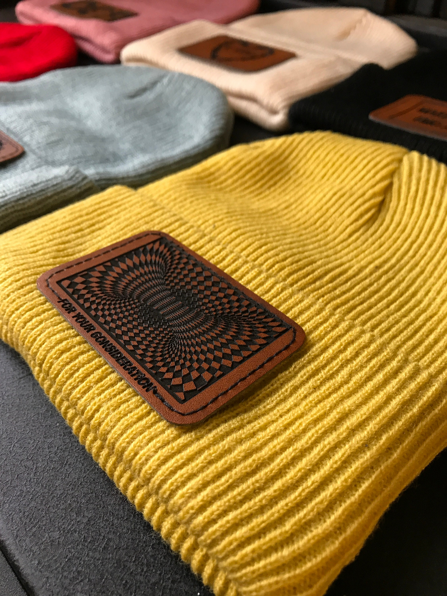 Yellow beanie with a brown patch featuring a geometric design, surrounded by other beanies in various colors.