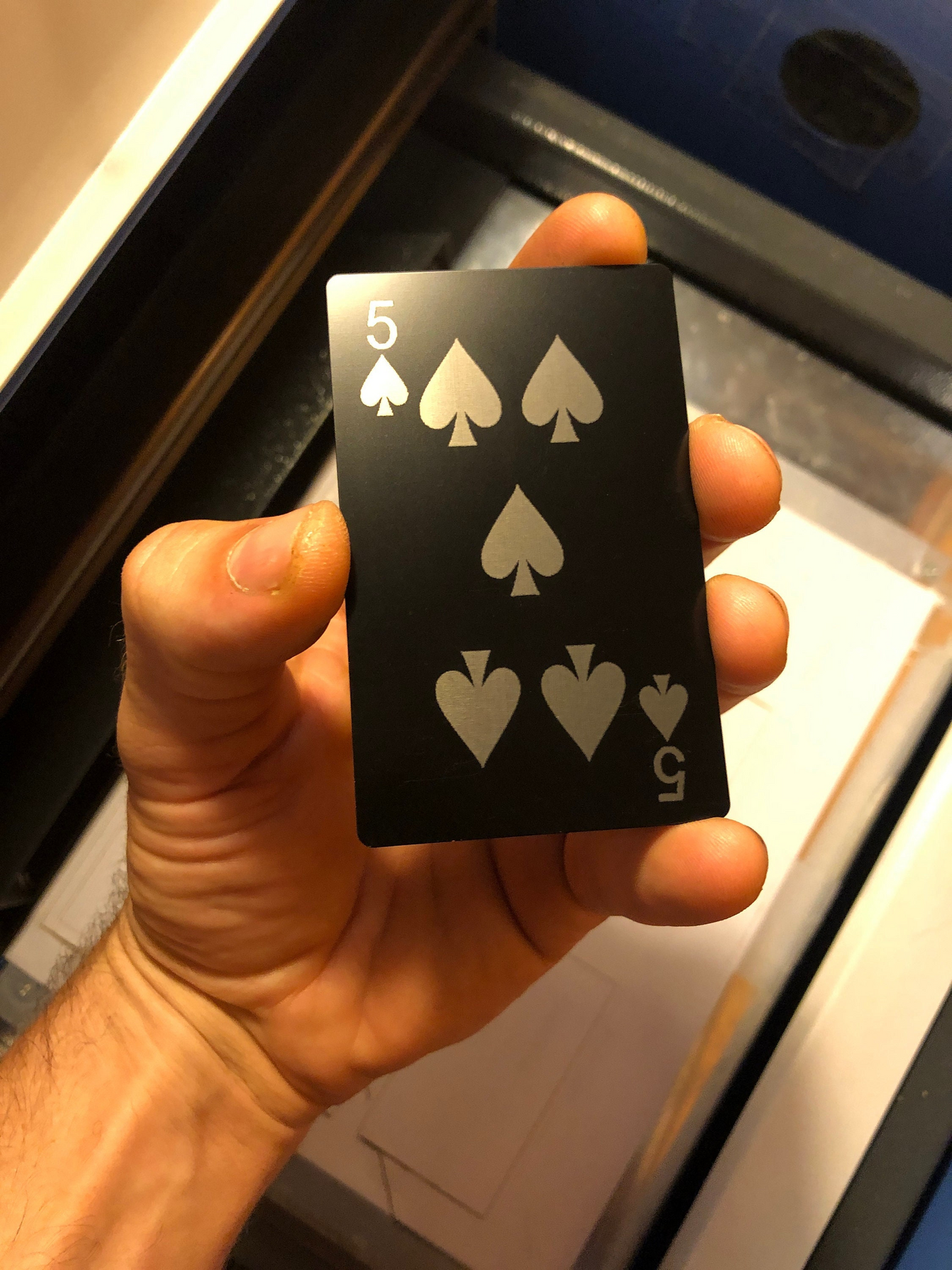 Hand holding a black metal poker card with five of spades design laser engraved in anodized aluminum