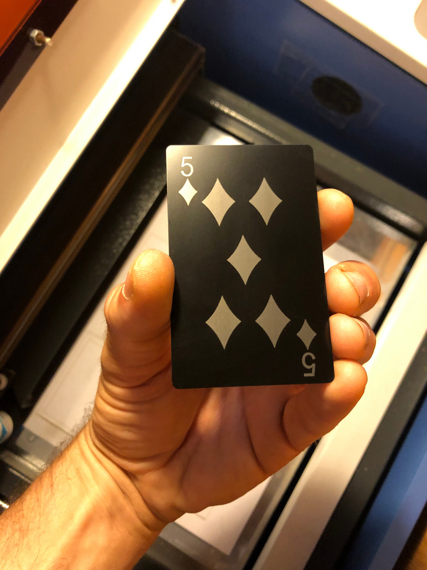 Hand holding a five of diamonds metal playing card in front of a CNC machine.