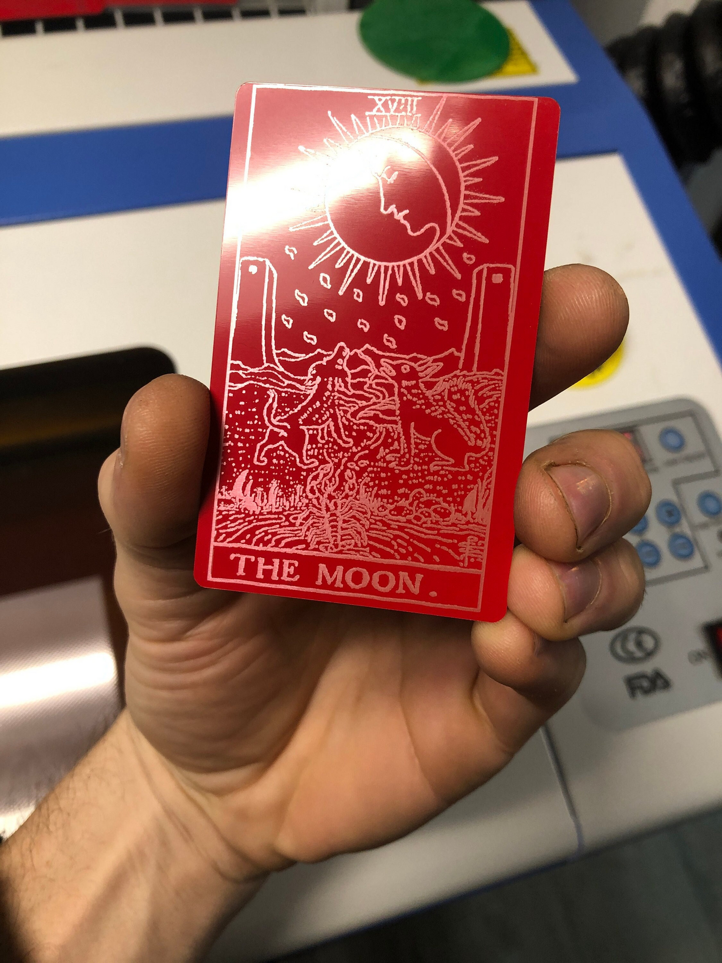 Red/silver metal tarot card titled 'The Moon' held by a hand with a machine in the background.