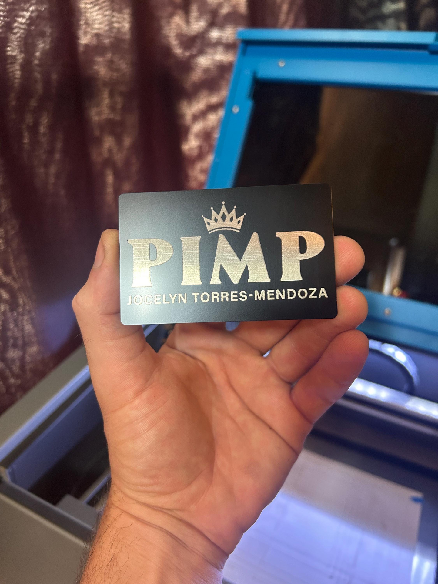 Hand holding a laser engraved metal keepsake gag gift card with 'PIMP' and a customized name printed on it, with a blurred background.