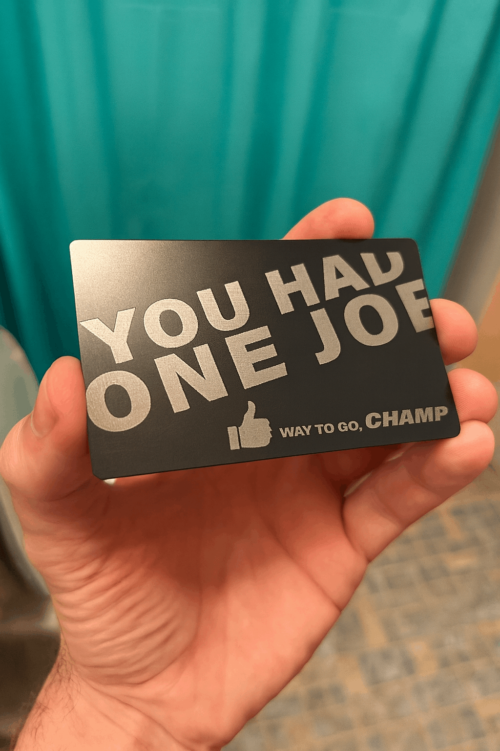 Hand holding a metal gag gift card with humorous You Had One Job text against a teal curtain background
