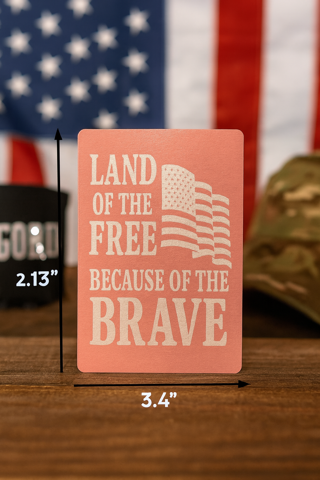Rectangular sign with 'Land of the Free Because of the Brave' text and American flag, displayed with measurements and an American flag and helmet in the background.