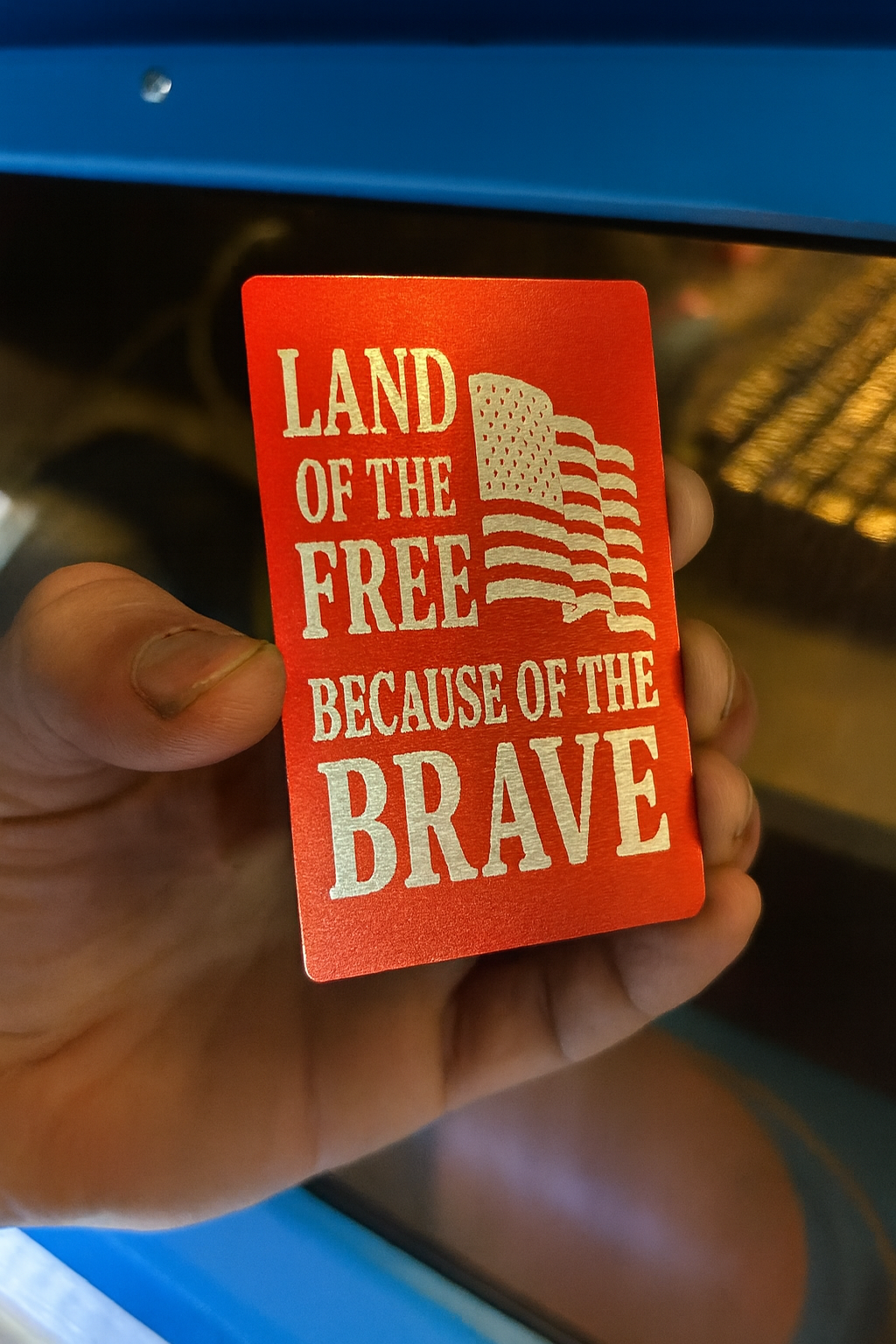 Red/silver metal card with 'Land of the Free Because of the Brave' text held by a hand.
