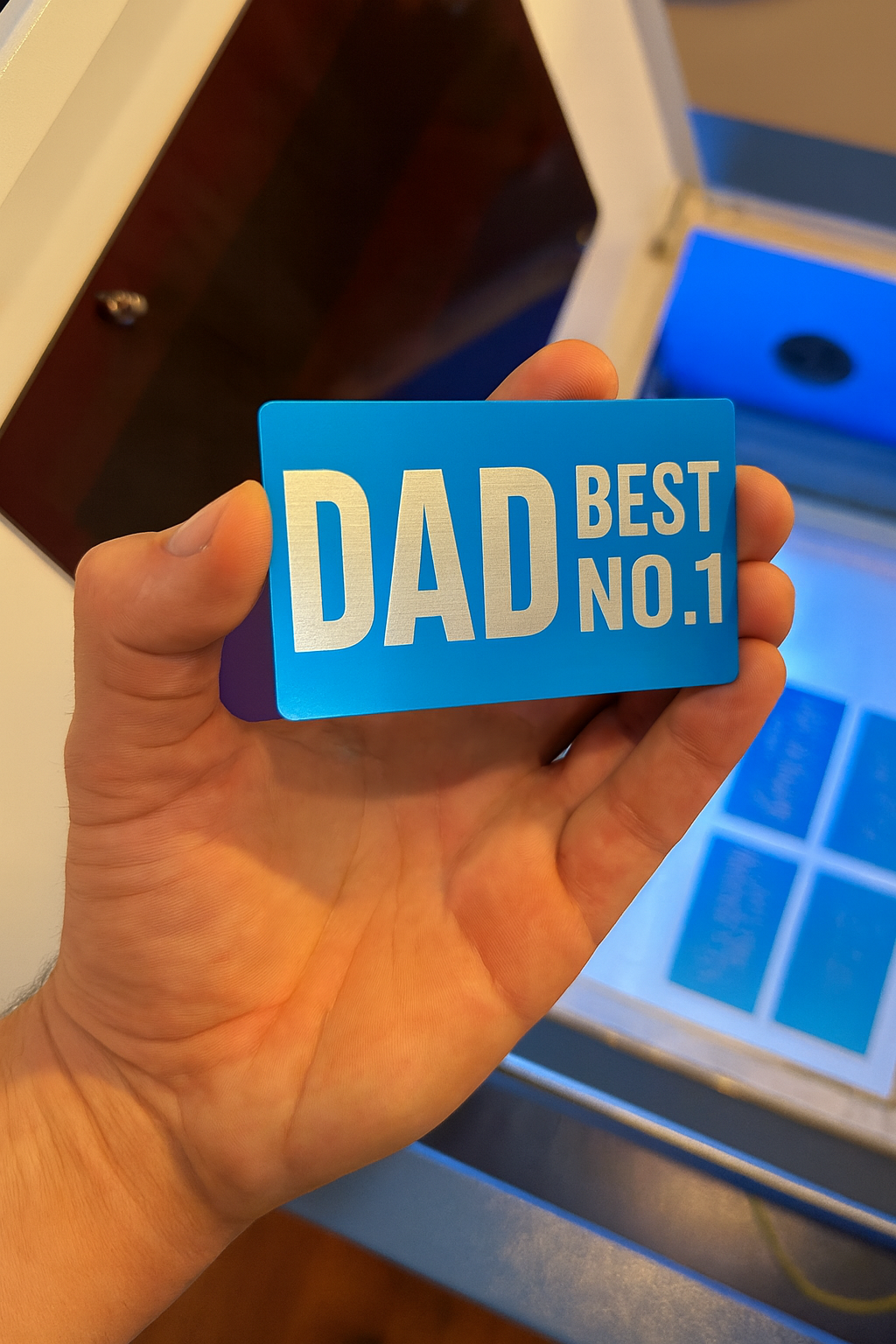 Hand holding a blue metal laser engraved card with 'DAD BEST NO.1' text in front of a blue and white background.