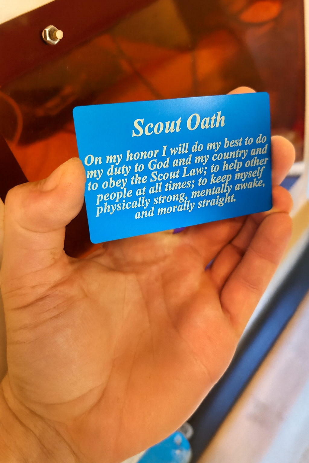 Hand holding a metal silver/blue card with 'Scout Oath' text engraved in anodized aluminum against a blurred background
