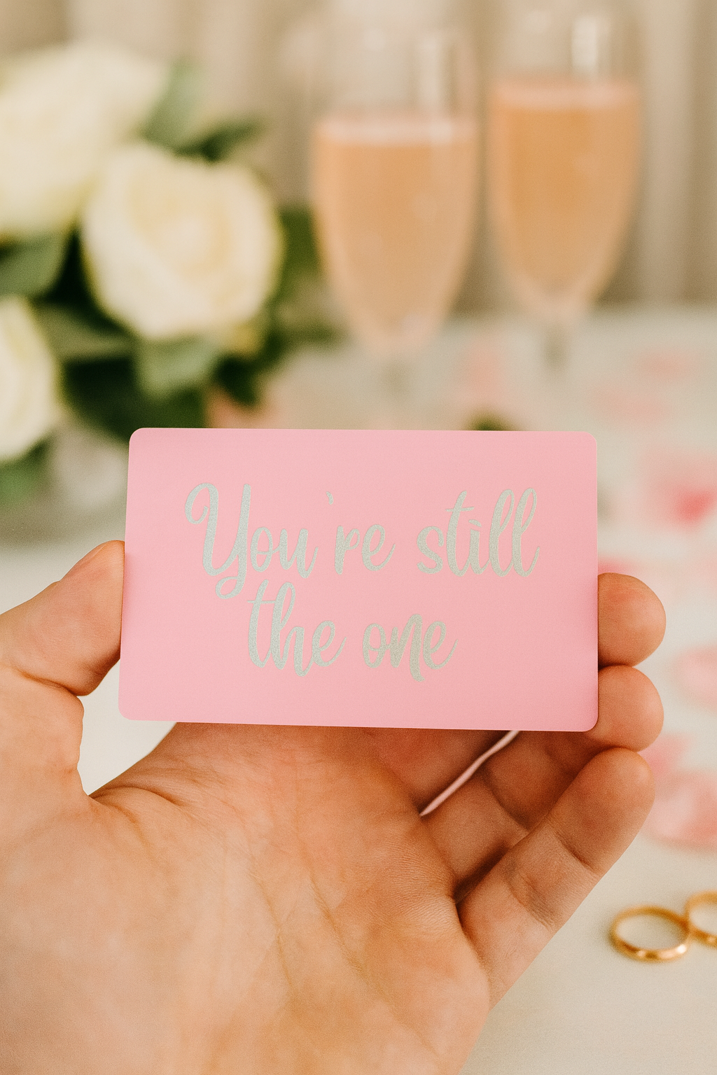 Hand holding a pink card with 'You're still the one' text, blurred background of flowers and glasses.