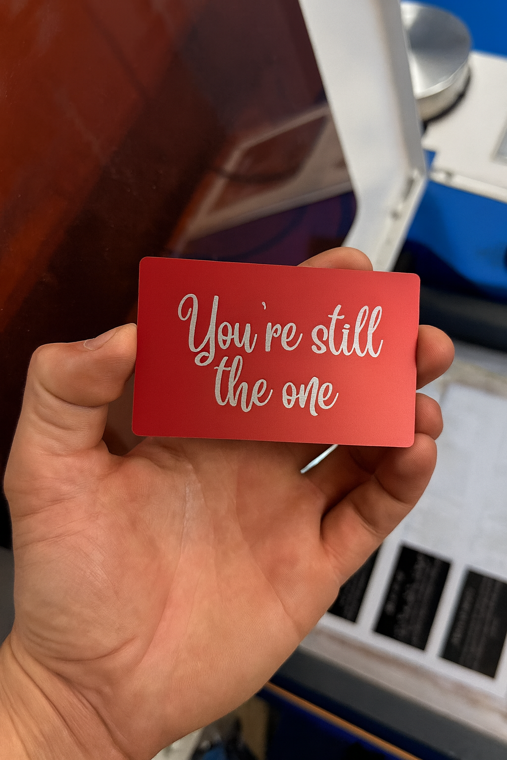 Hand holding a red/silver anodized aluminum romantic gift card with 'You're still the one' text laser engraved, presented in a blurred indoor setting