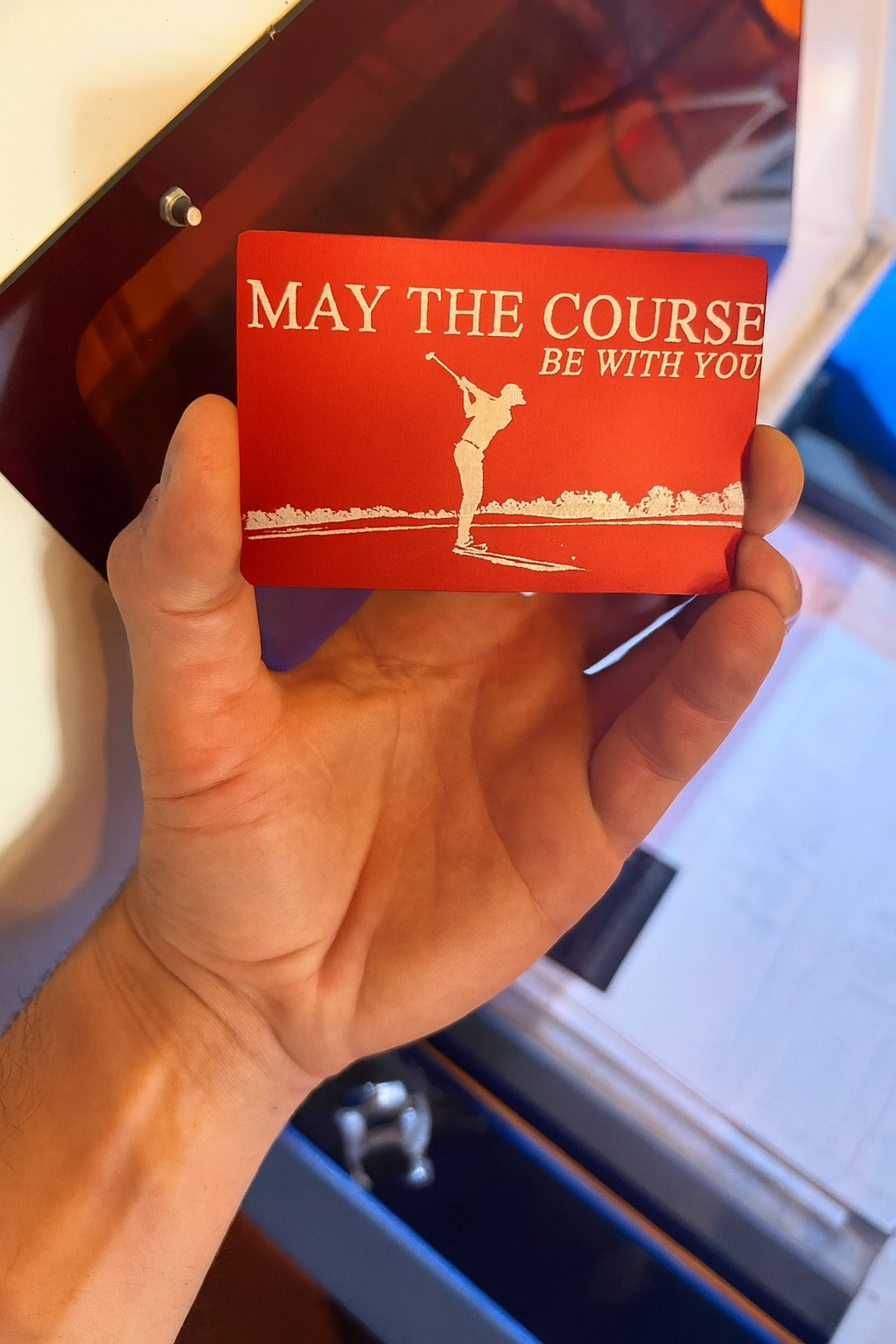 Hand holding a red/silver metal card with a golf design and inspirational text, in an industrial machine setting