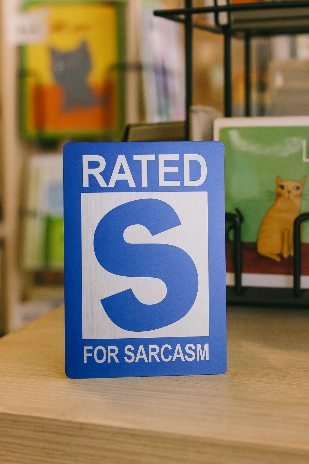 Blue/silver mini novelty metal sign with 'Rated S for Sarcasm' text on a wooden surface.