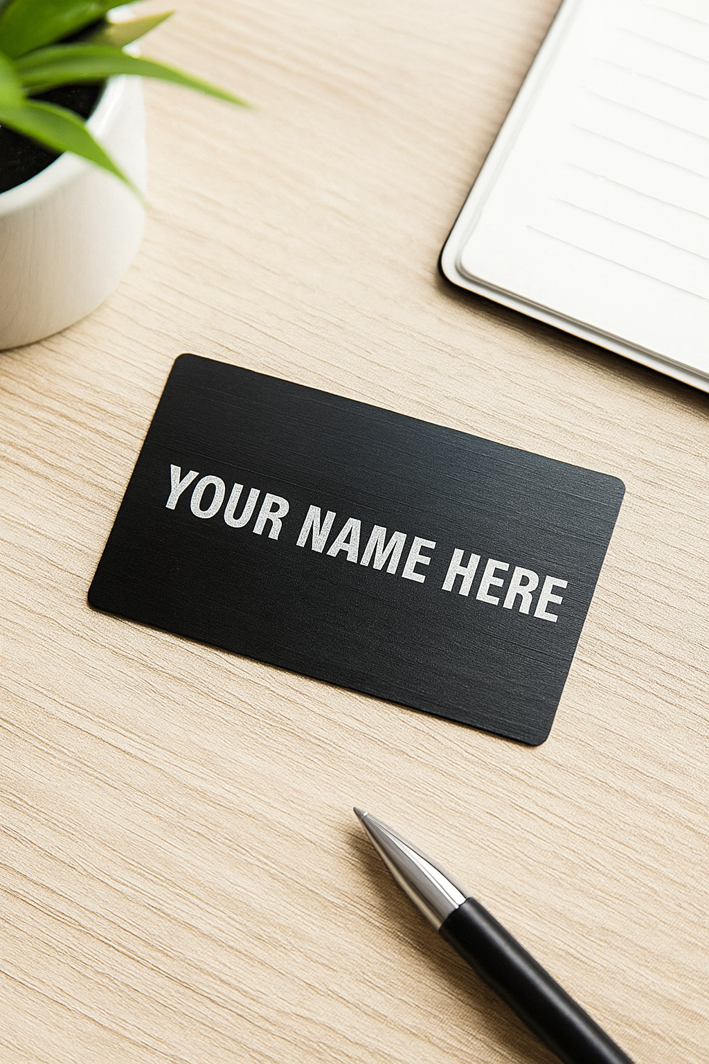 Black metal business card with 'YOUR NAME HERE' example text on a wooden desk with a pen and plant.