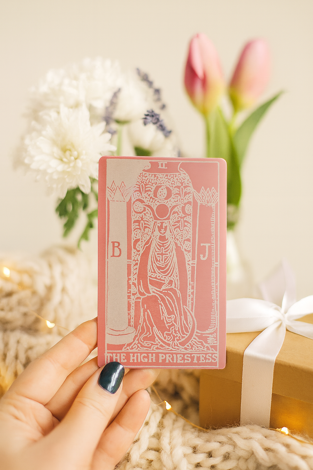 Rose gold metal tarot card held by a hand with flowers and gift in the background