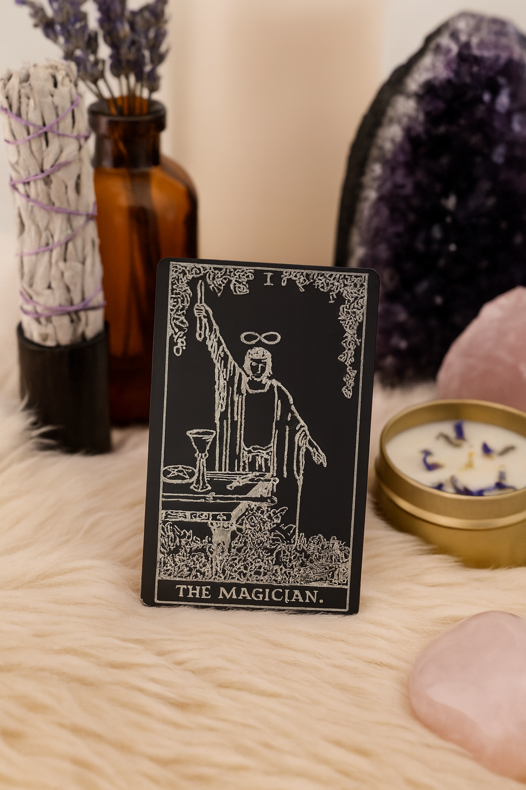 Metal tarot card titled 'The Magician' surrounded by mystical items like a crystal and candles on a soft surface.
