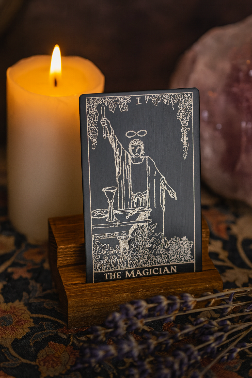 Metal Tarot card in black/silver titled 'The Magician' on a stand with a lit candle and lavender flowers in the background.
