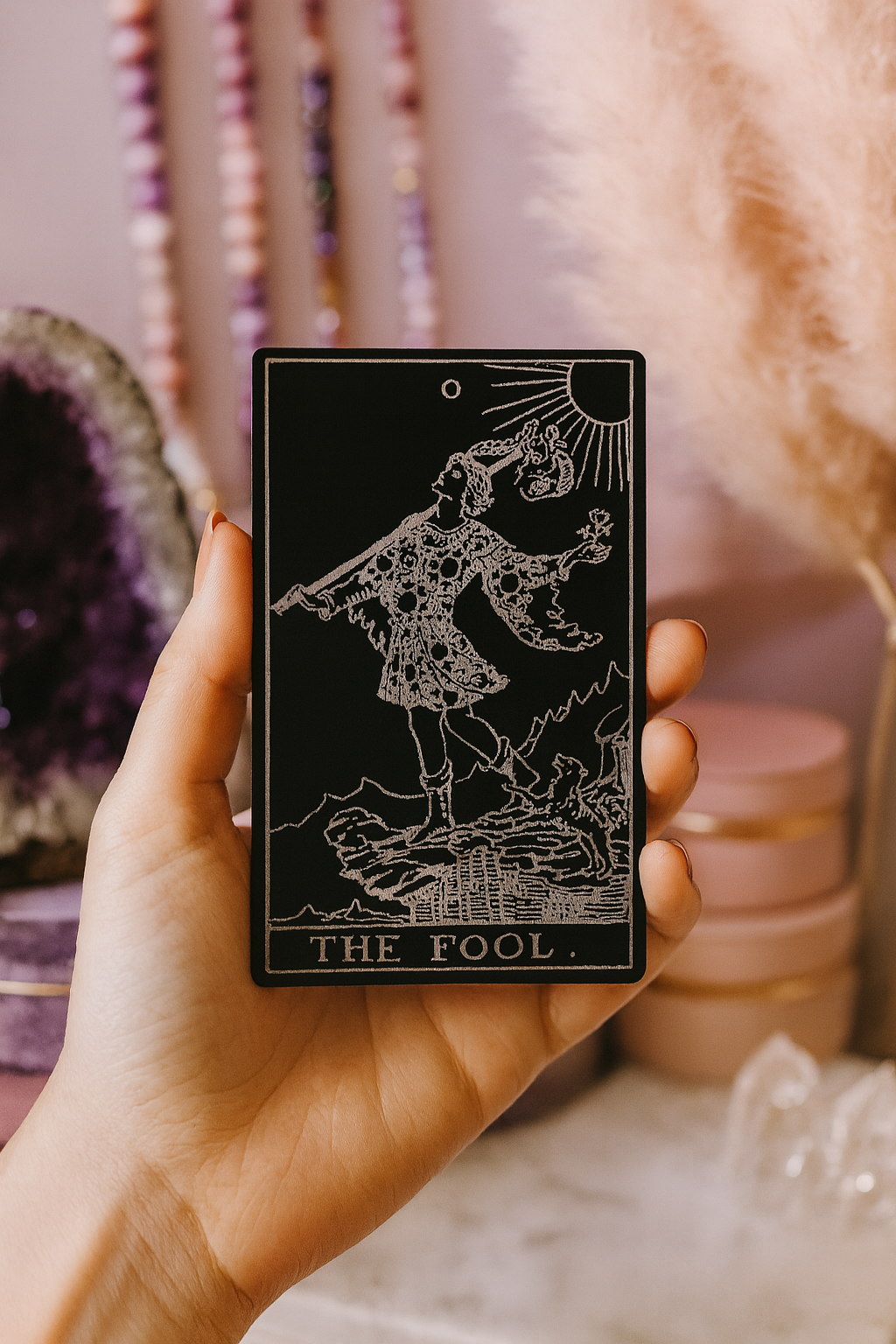 Hand holding a laser engraved metal tarot card with 'The Fool' illustration against a mystical background.