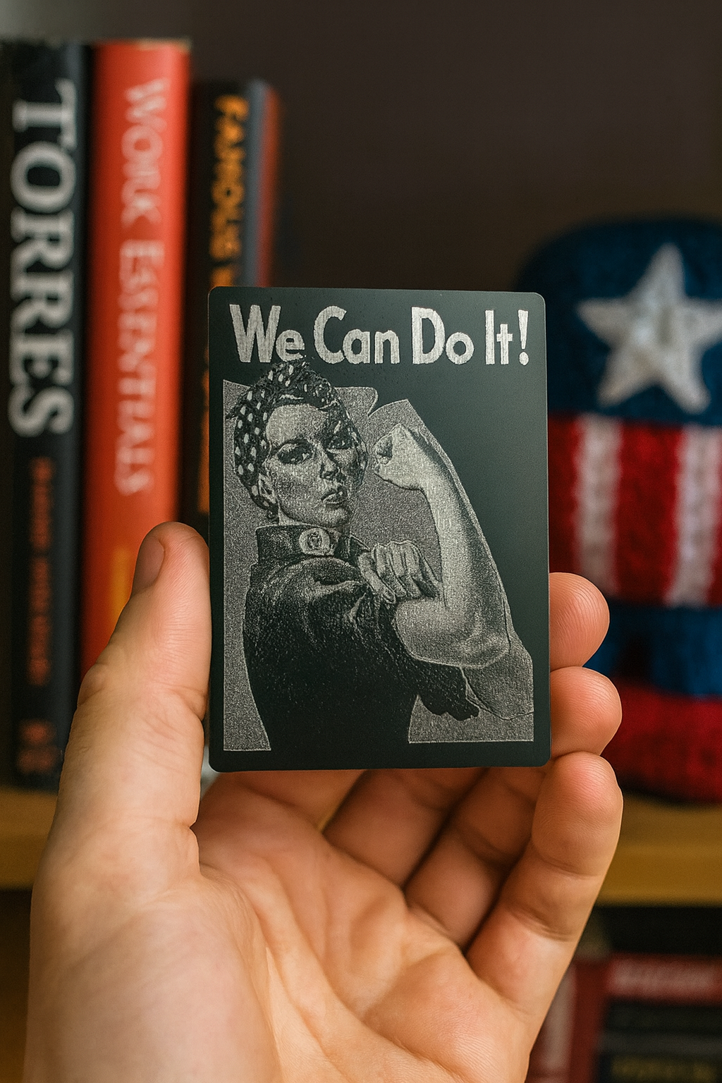 Hand holding a metal laser engraved inspirational card with 'We Can Do It!' text and a Rosie the Riveter graphic, with books and Americna art in the background.