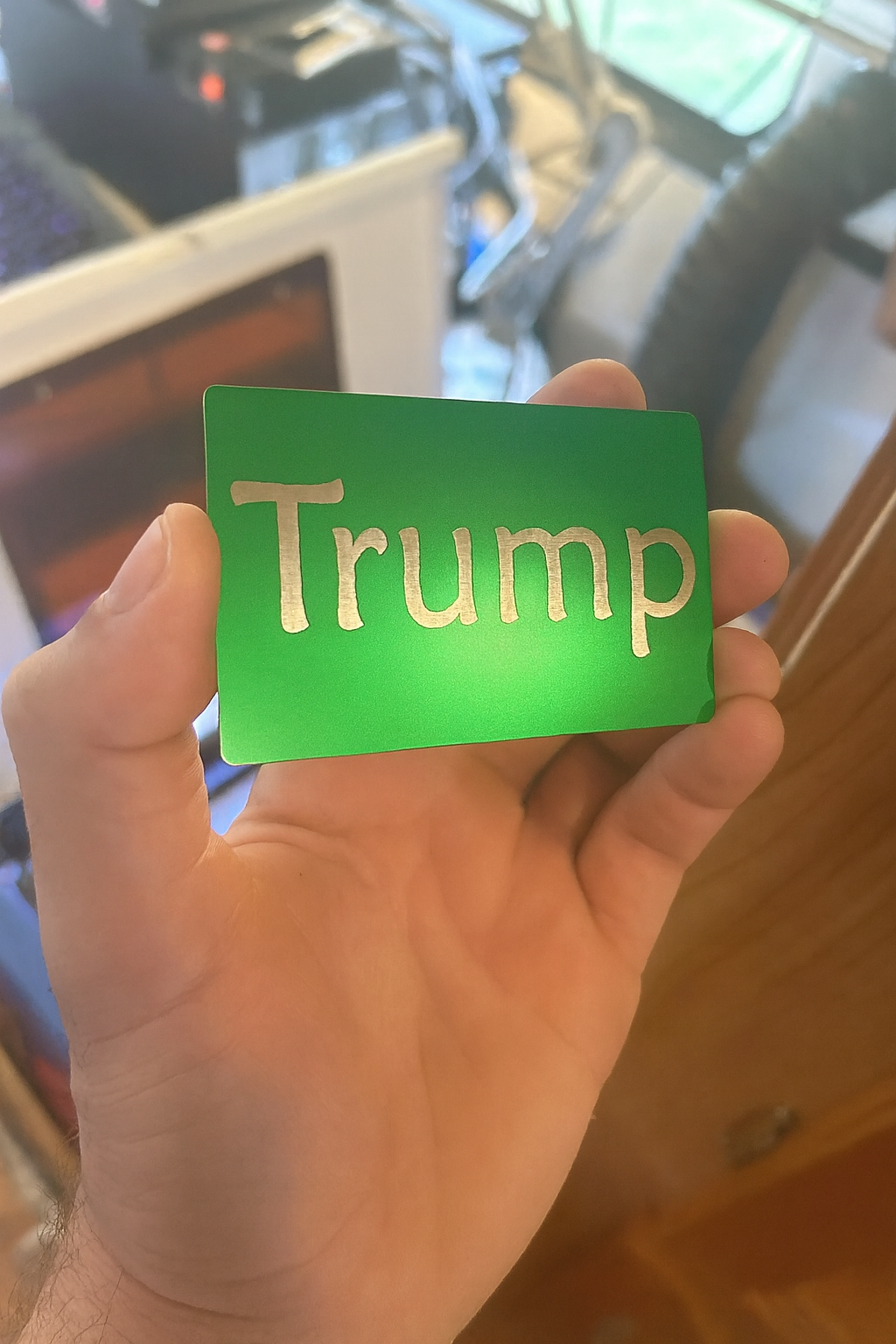Hand holding a green/silver metal card with 'Trump' laser engraved on it, inside a laser engraving cnc shop.