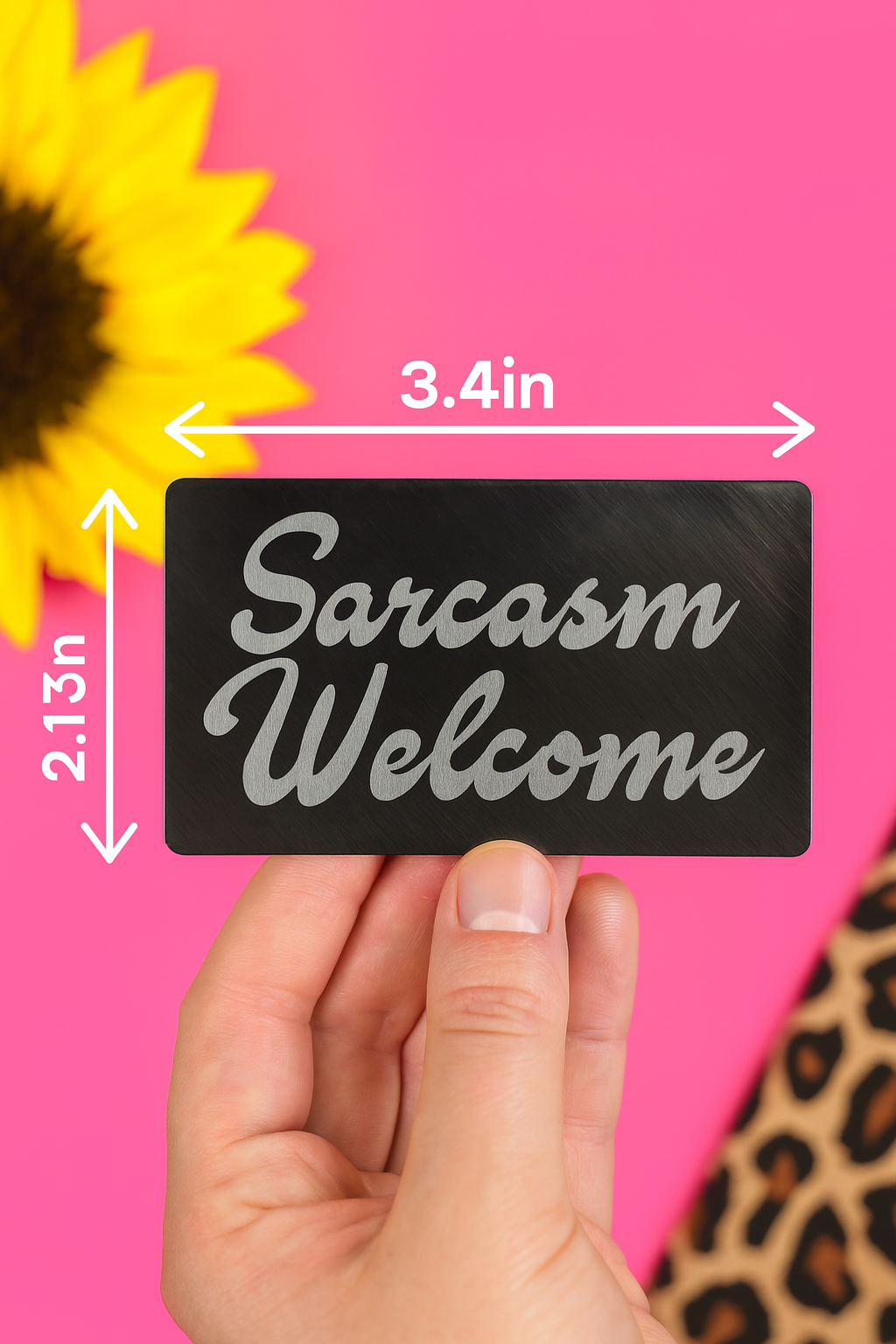 Hand holding a sign with 'Sarcasm Welcome' text with size/dimensions against a pink background with a sunflower.