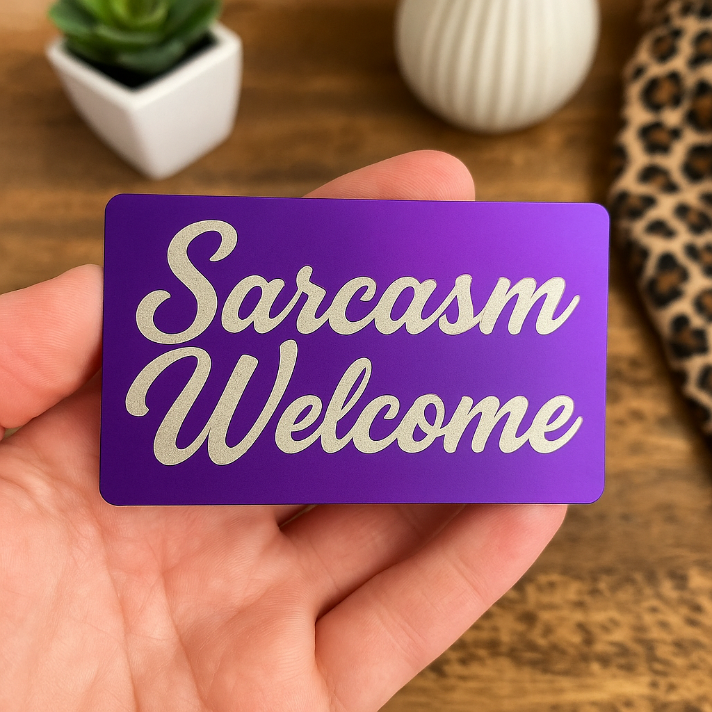 Hand holding a purple/silver metal card with 'Sarcasm Welcome' text on a wooden surface.