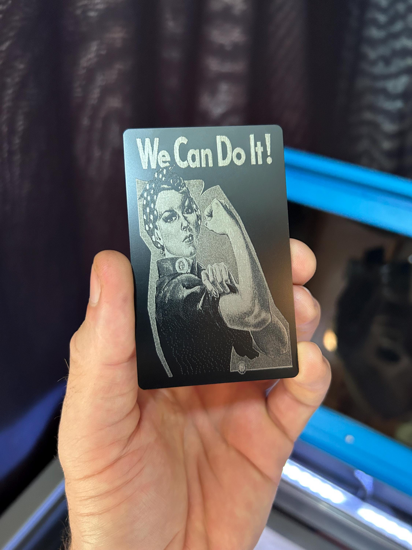 Hand holding a laser engraved metal keepsake card with 'We Can Do It!' and a Rosie the Riveter graphic in matte black/silver