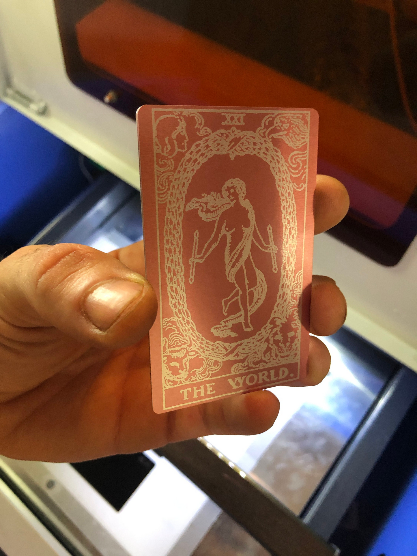 Hand holding a pink (rose gold) and silver laser engraved metal tarot card with intricate design and text.