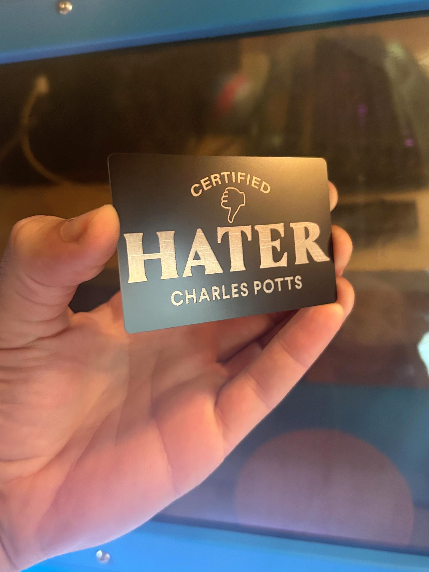 Hand holding a card labeled 'Certified Hater' with a personalized name engraving