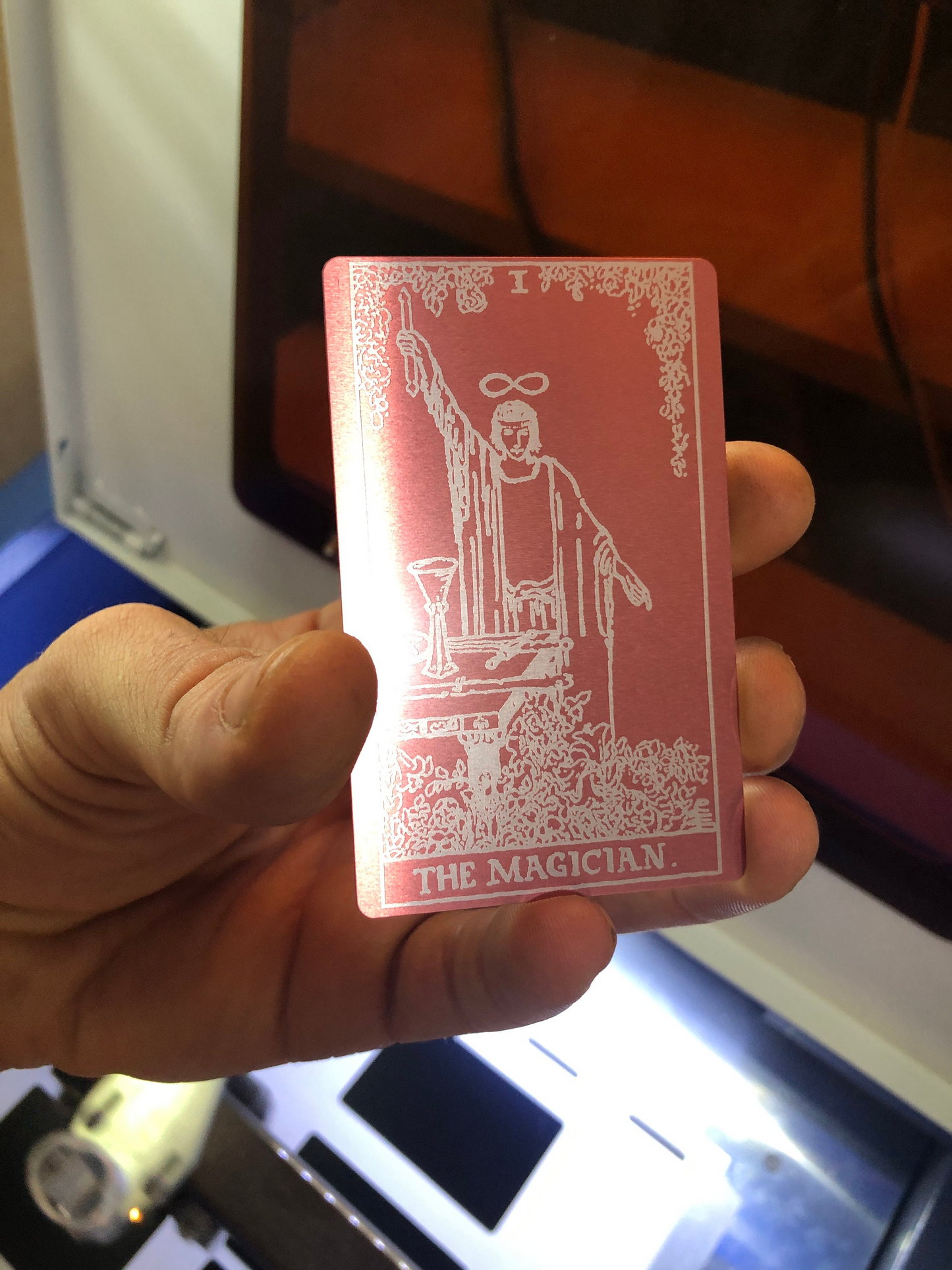 Hand holding a rose gold (pink)/silver laser engraved metal tarot card with white illustrations and text, featuring 'The Magician' from a tarot deck.