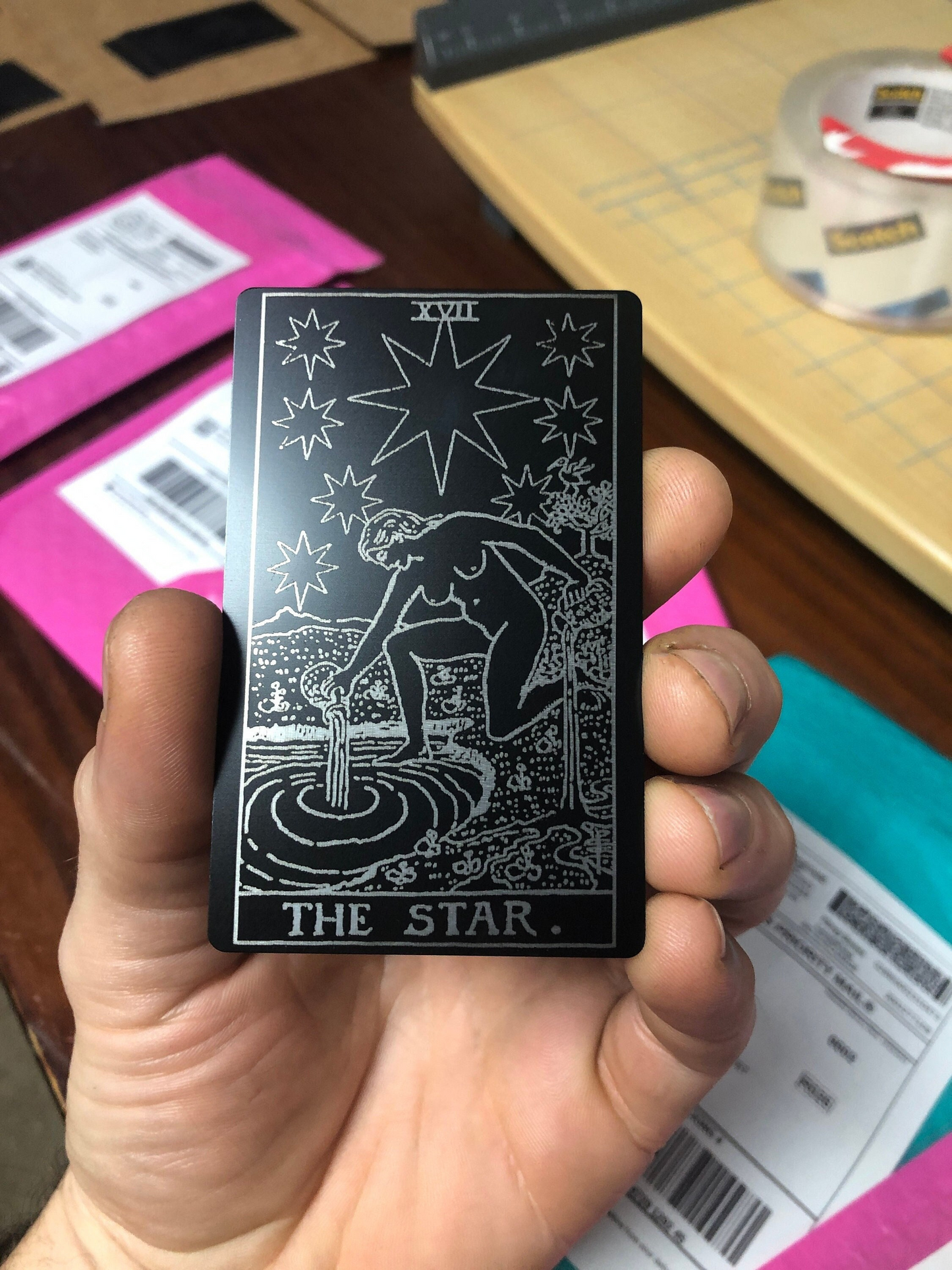 Hand holding a metal laser engraved tarot card titled 'The Star' with a star and person illustration, on a blurred background.