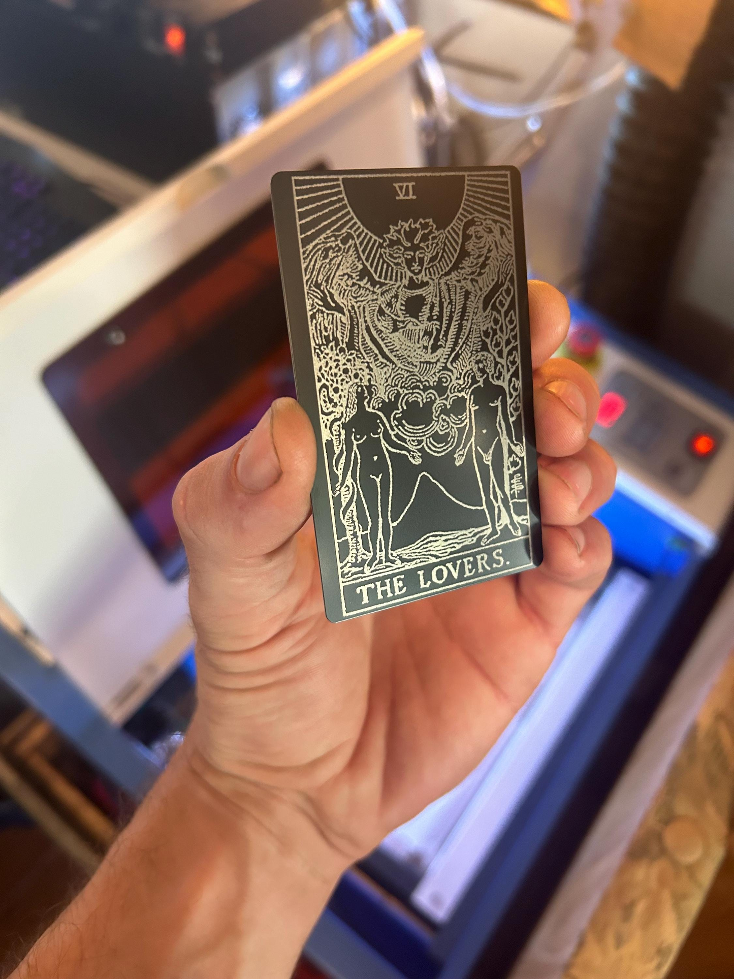Hand holding a metal laser engraved tarot card titled 'The Lovers' in front of a blurred background.