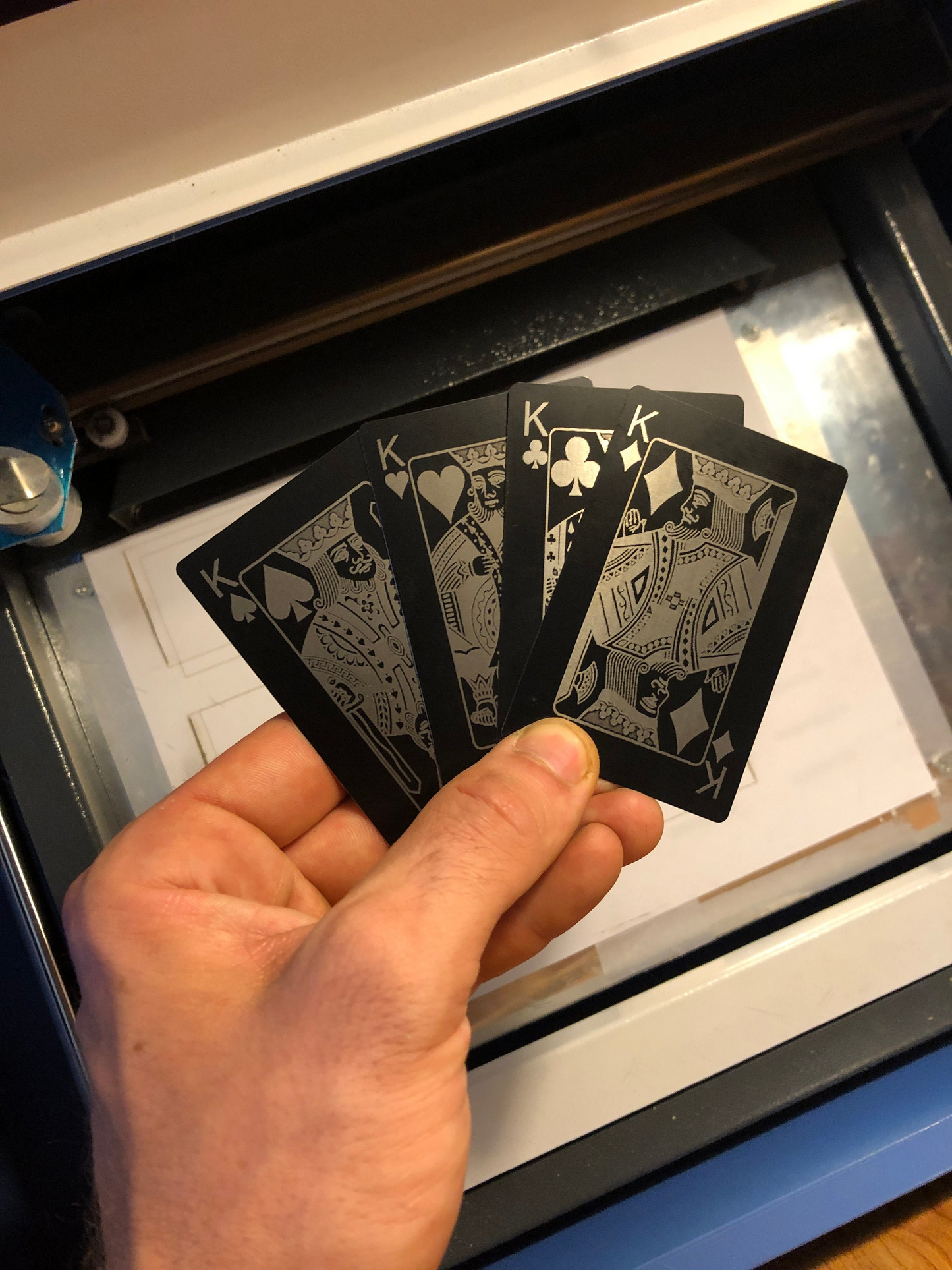 Hand holding four kings (spades, hearts, clubs, diamonds) in black/silver metal playing cards with a machine in the background