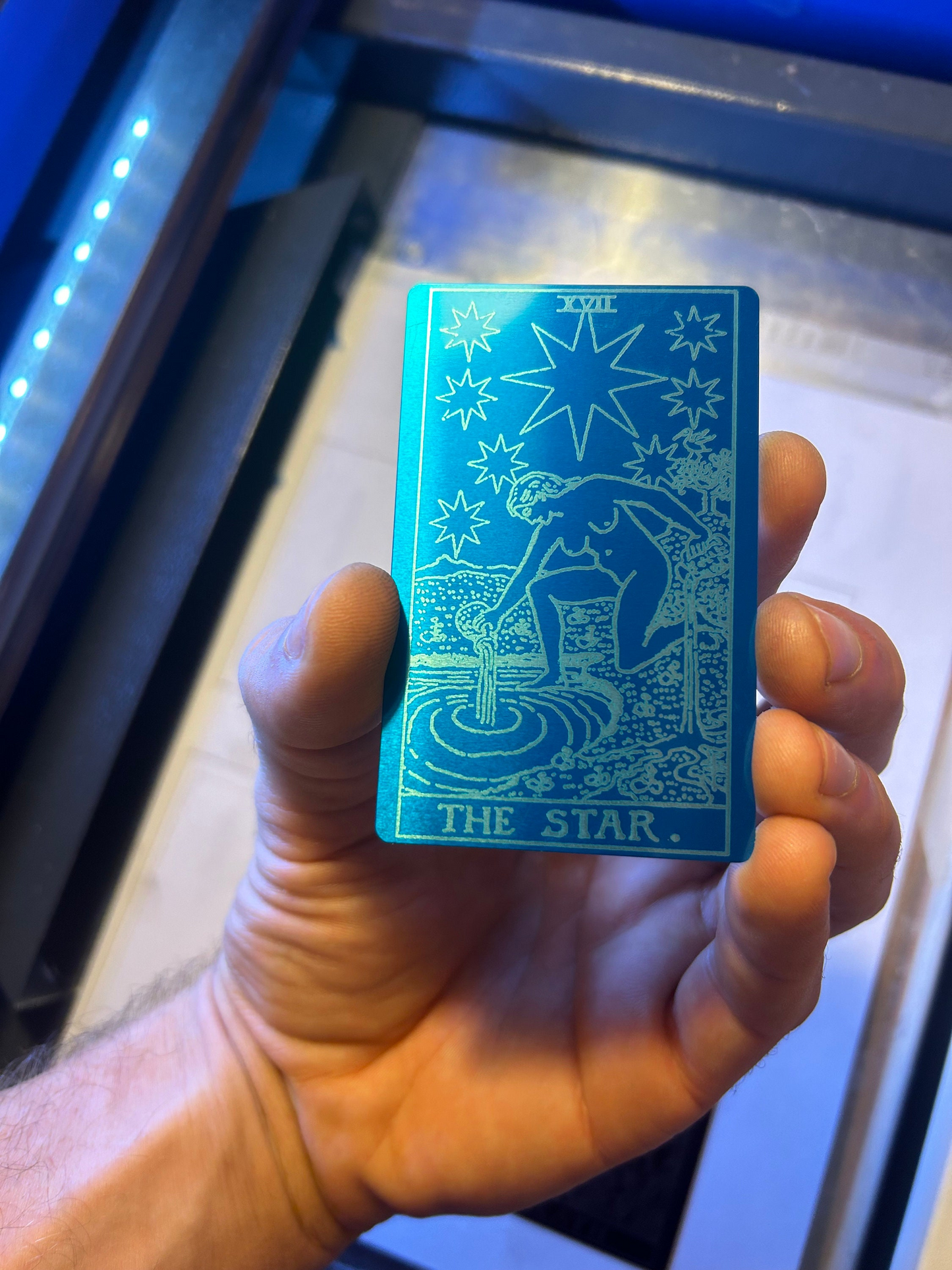 Hand holding a laser engraved anodized aluminum blue/silver tarot card titled 'The Star' against a reflective surface.