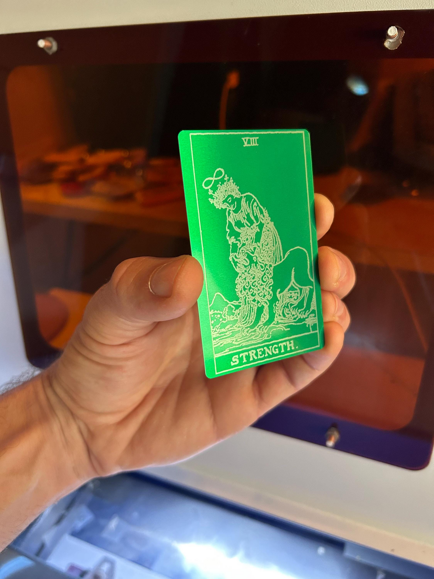 Hand holding a laser engraved metal green/silver anodized aluminum tarot card with Strength design against a blurred background