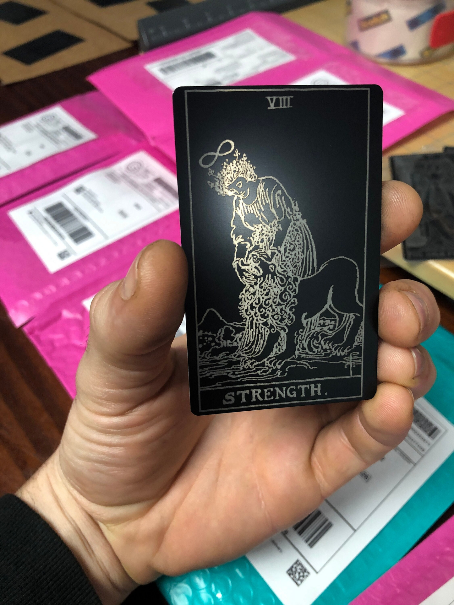 Hand holding a metal laser engraved tarot card with 'Strength' and a lion design, with a pink box in the background.
